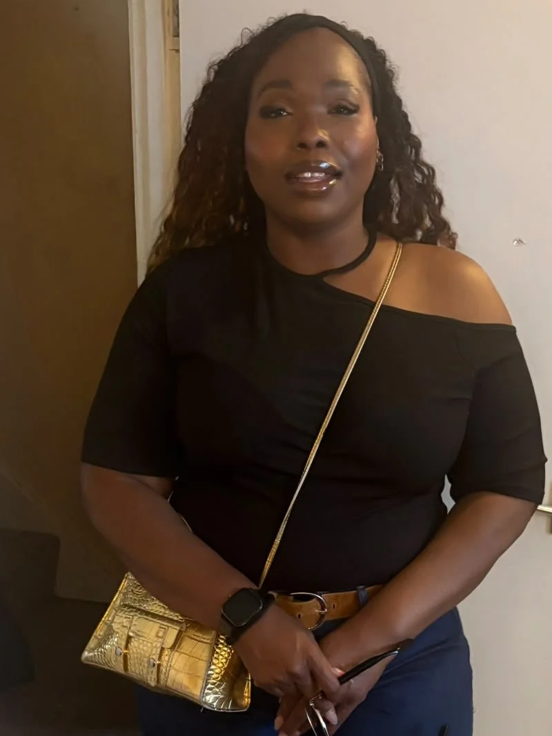 A woman with dark curly hair wearing a black off-the-shoulder top, accessorized with a gold chain strap purse, a black watch, and earrings, standing indoors in front of a plain wall.