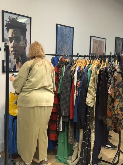 A person with blonde hair wearing a beige outfit shopping at a clothing store, looking at a rack of various colorful clothes, with framed artwork on the wall in the background.