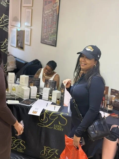 Woman in a black jacket and baseball cap with a Lakers logo at a perfume booth, smiling at the camera. Behind her are other people and perfume displays.