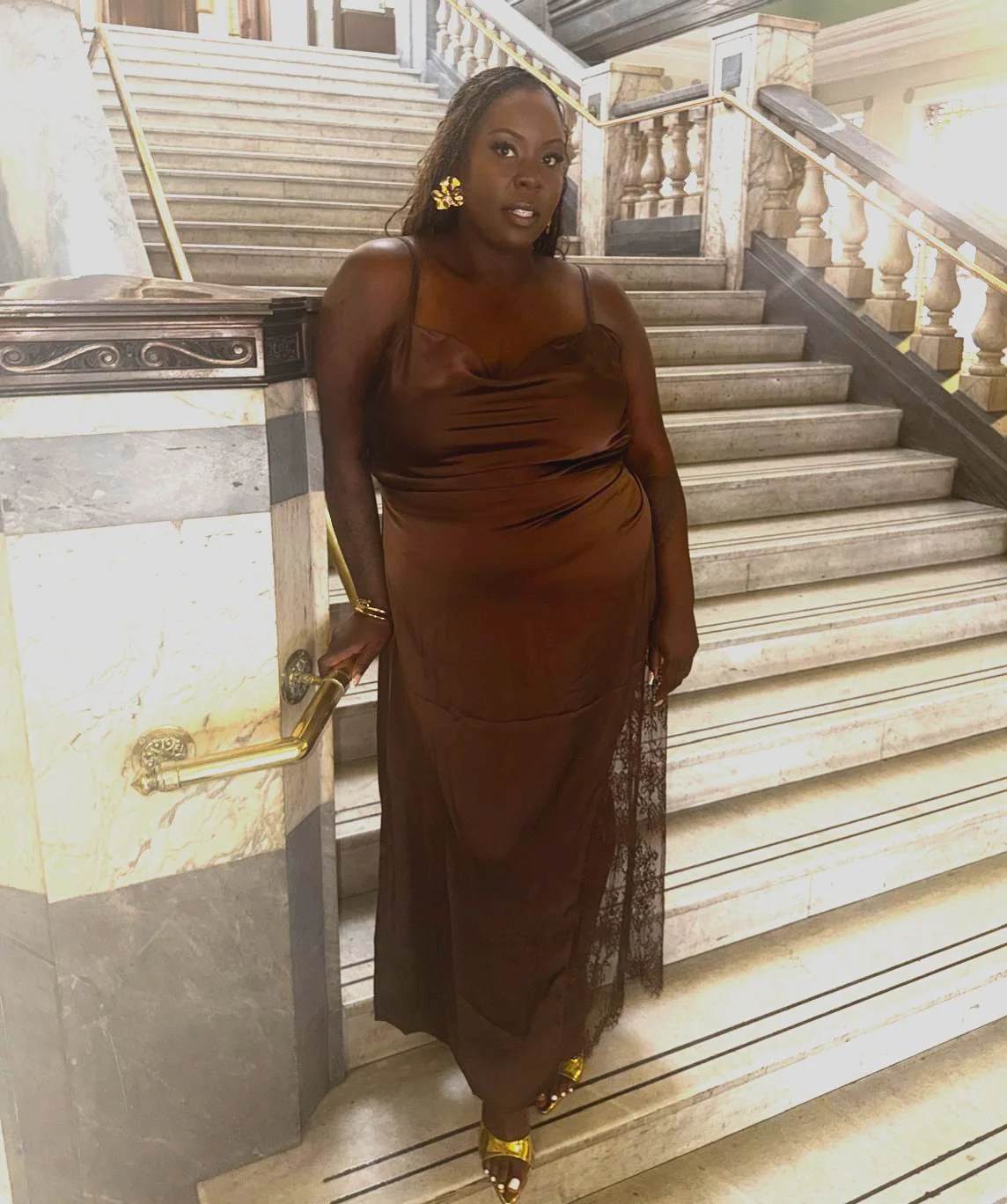 A woman in a brown gown standing on marble stairs inside a building with ornate railings and marble decor.