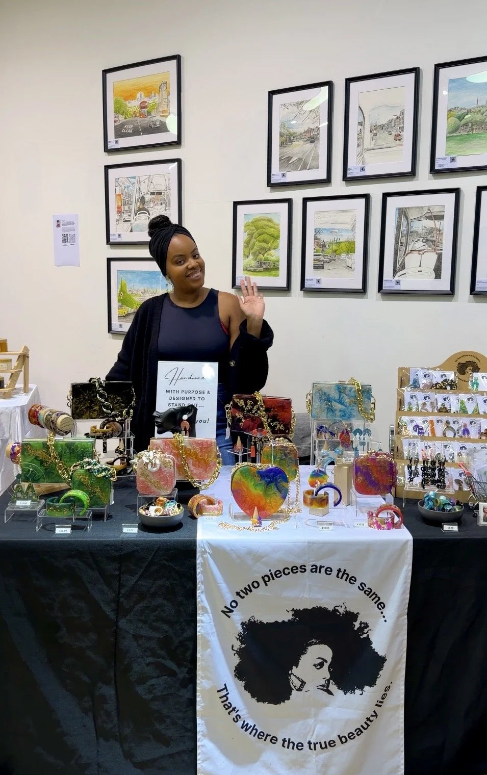 A woman standing behind a table displaying colorful handmade jewelry and accessories at an art fair or market, with framed landscape artwork on the wall behind her.