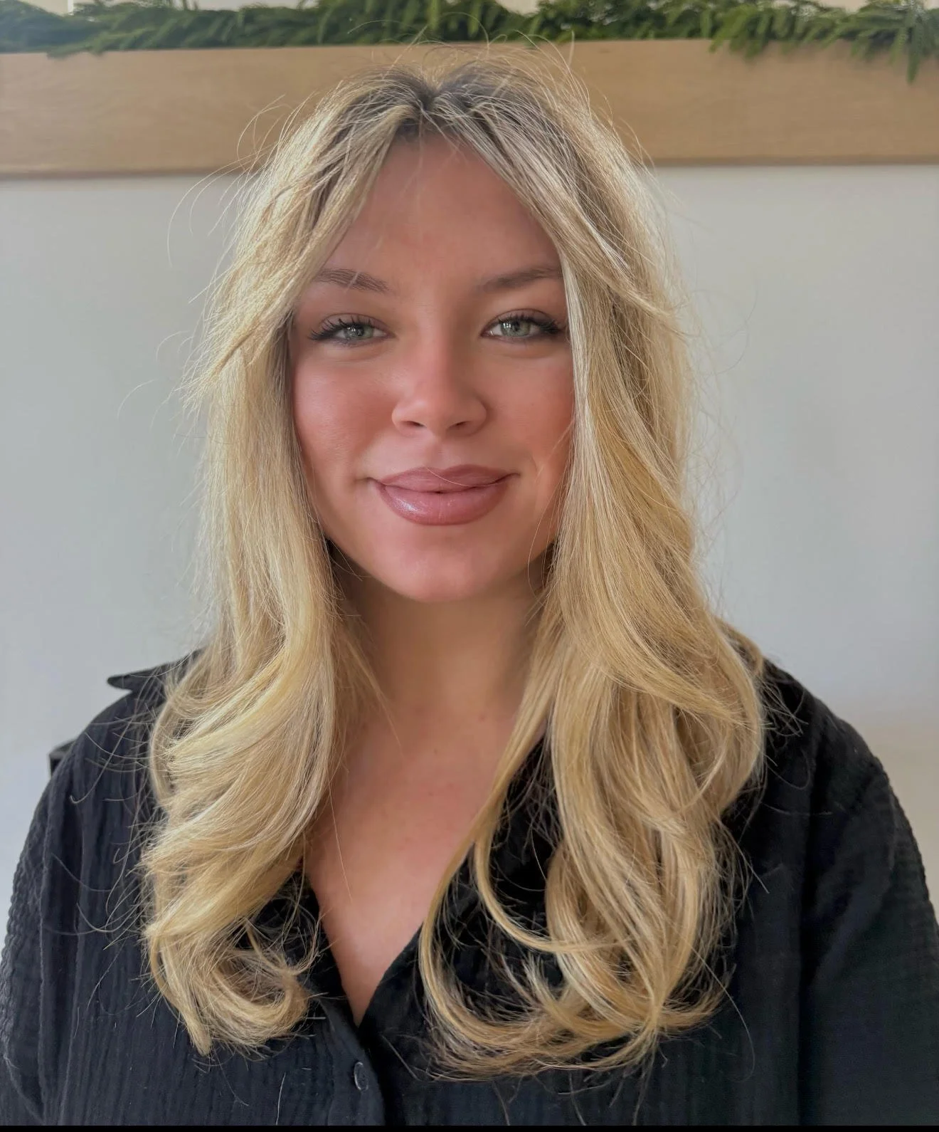 A woman with long, wavy blonde hair and blue eyes, wearing a black top, smiling at the camera. Behind her is a plain wall and a wooden piece of decor with green plants.