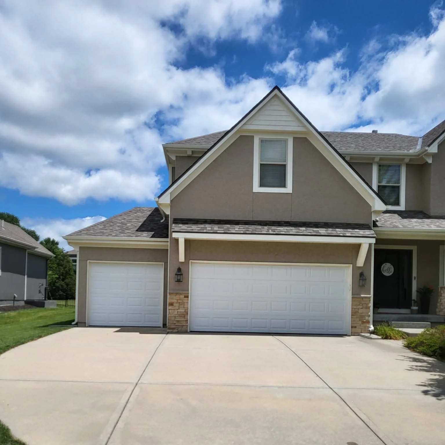 Front view of a modern two-story house with two garage doors, a driveway, and a small front yard. The house has beige walls, white trim, and a dark gray roof, under a partly cloudy sky.