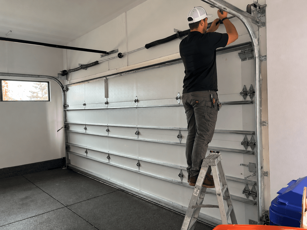 A man standing on a step ladder installing or repairing a sectional garage door in a garage. He is wearing a black shirt, gray pants, and a white cap.
