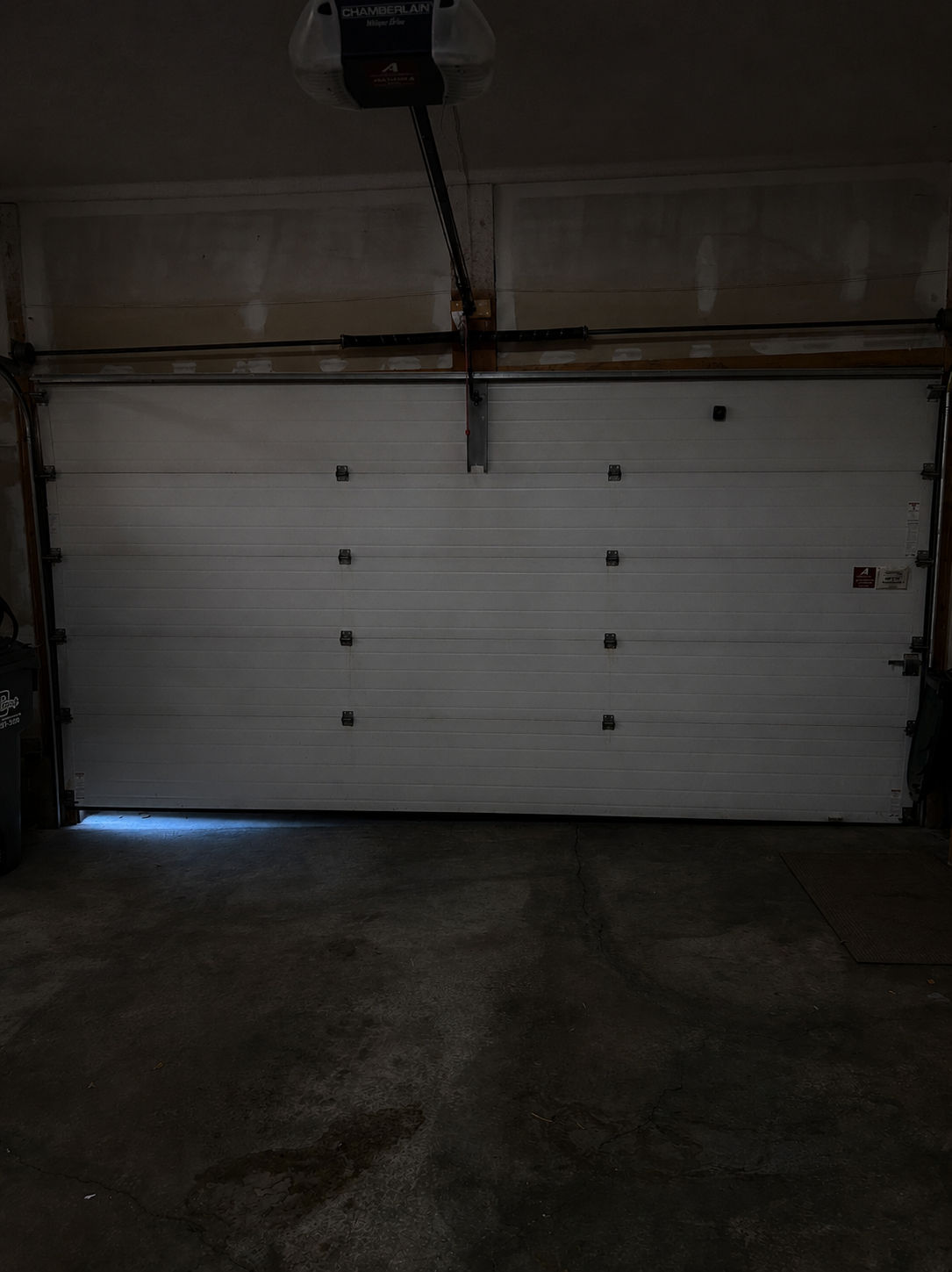 White sectional garage door with metal hardware, closed in a dimly lit garage, with faint light coming from a small gap at the bottom.
