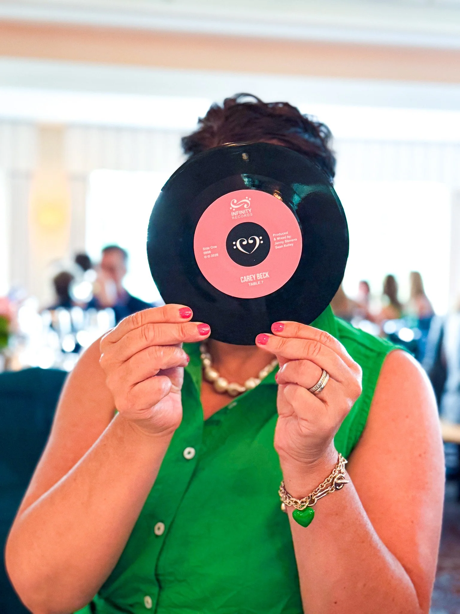 Woman in a green dress holding a black vinyl record with a pink label in front of her face. The record's label reads 'Infinity Records' and 'Carey Beck.'