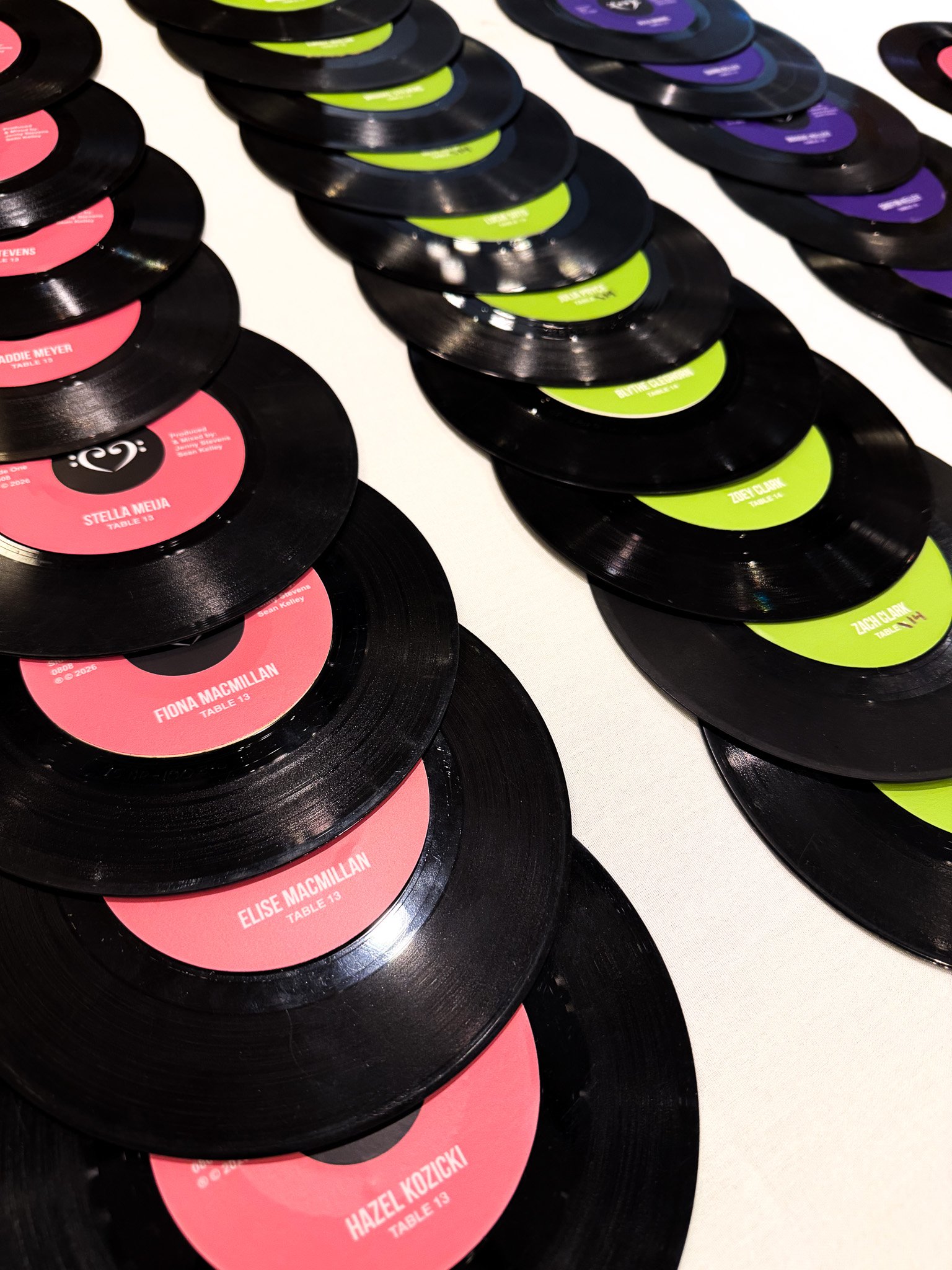 Stacks of black vinyl records with colorful labels laid out on a white surface. The labels are pink, green, and purple with text indicating the names of people and table numbers.