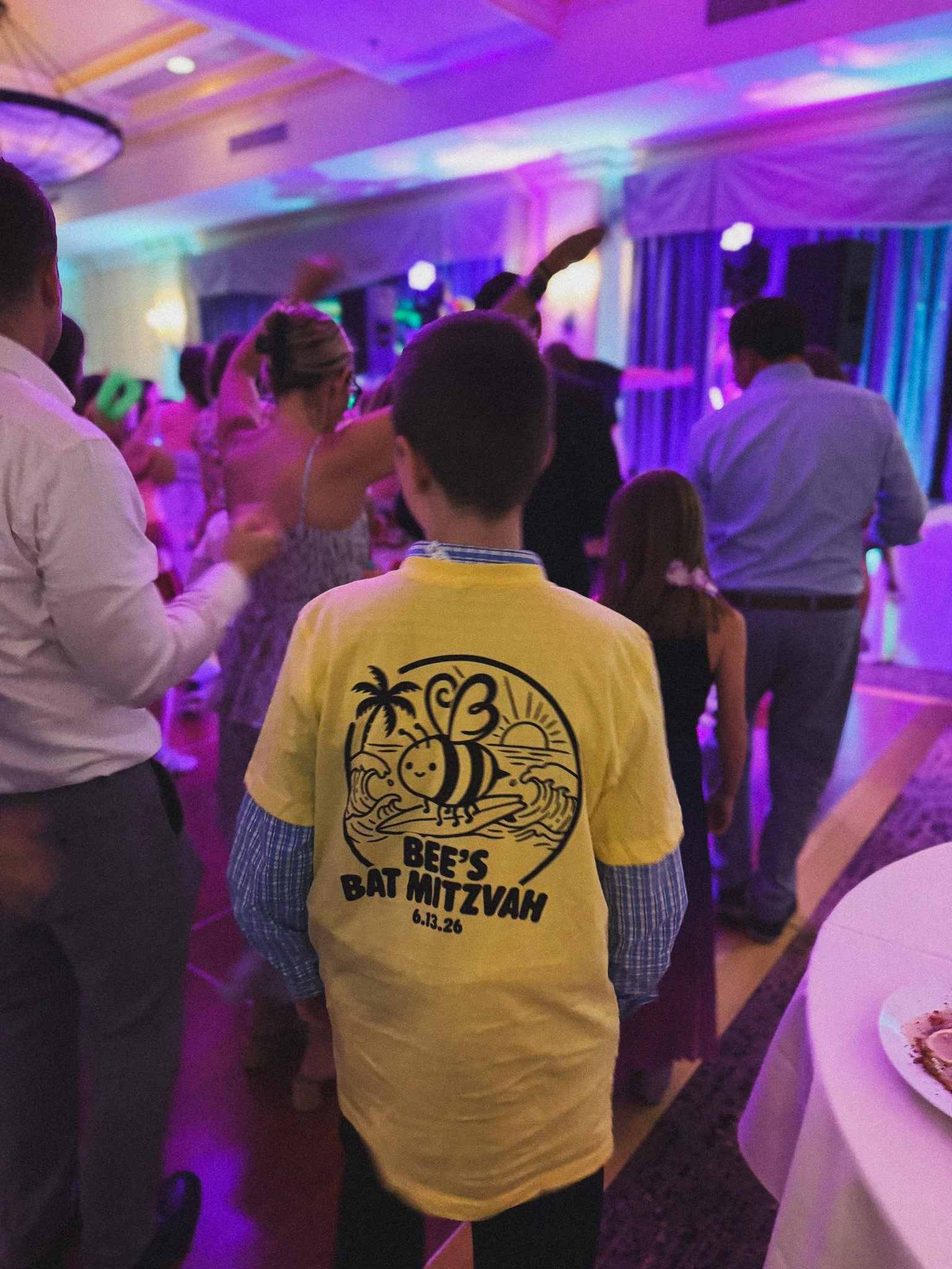 A group of people gathered indoors under colorful purple and pink lighting for a celebration event. A person in the foreground is wearing a yellow T-shirt with a bee and palm tree graphic, and text reading 'Bee's Bat Mitzvah 6.13.26.' The scene suggests a lively and festive atmosphere.
