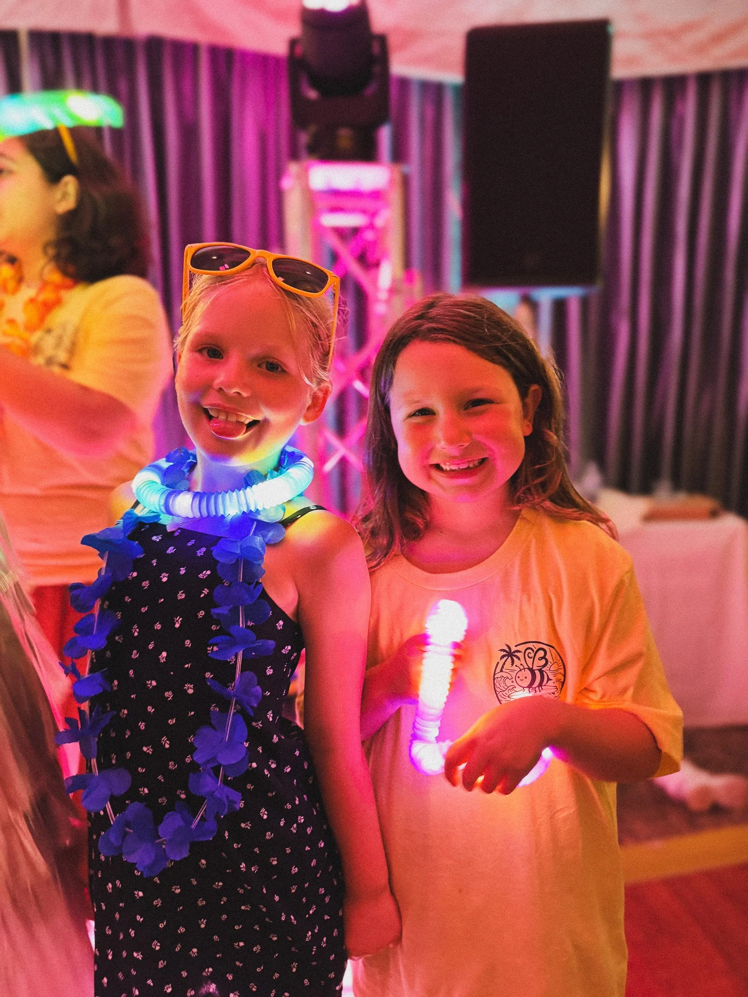 Two children smiling at a party, with one holding a glowing toy and the other wearing sunglasses on her head, standing under colorful lighting with a DJ booth and speakers in the background.