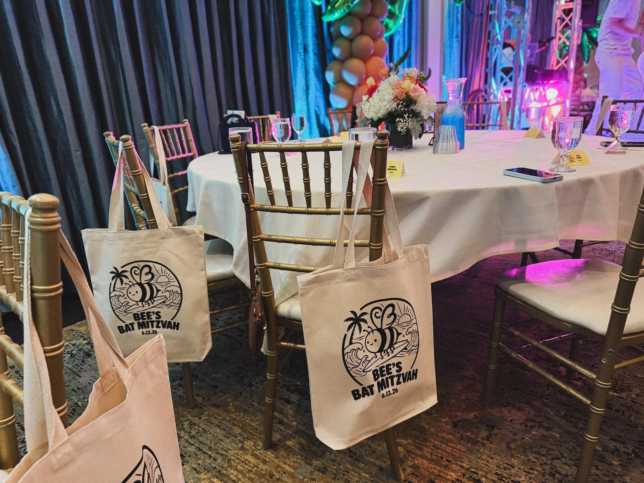 A banquet table decorated with floral centerpiece, water glasses, and table numbers, with golden chairs and beige tote bags hanging from some chairs; the event appears lively with colorful lighting and drapery, and features a bee-themed decoration that reads 'Bee's Bat Mitzvah 6.13.26'.
