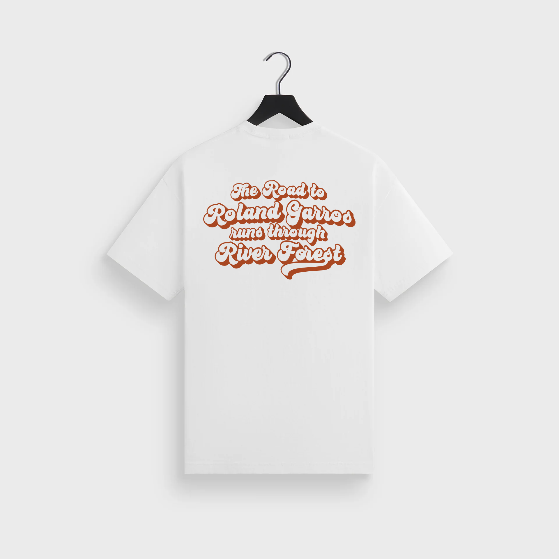 White T-shirt hanging on a black hanger with text 'The Road to Roland Garros runs through River Forest' printed in brown.