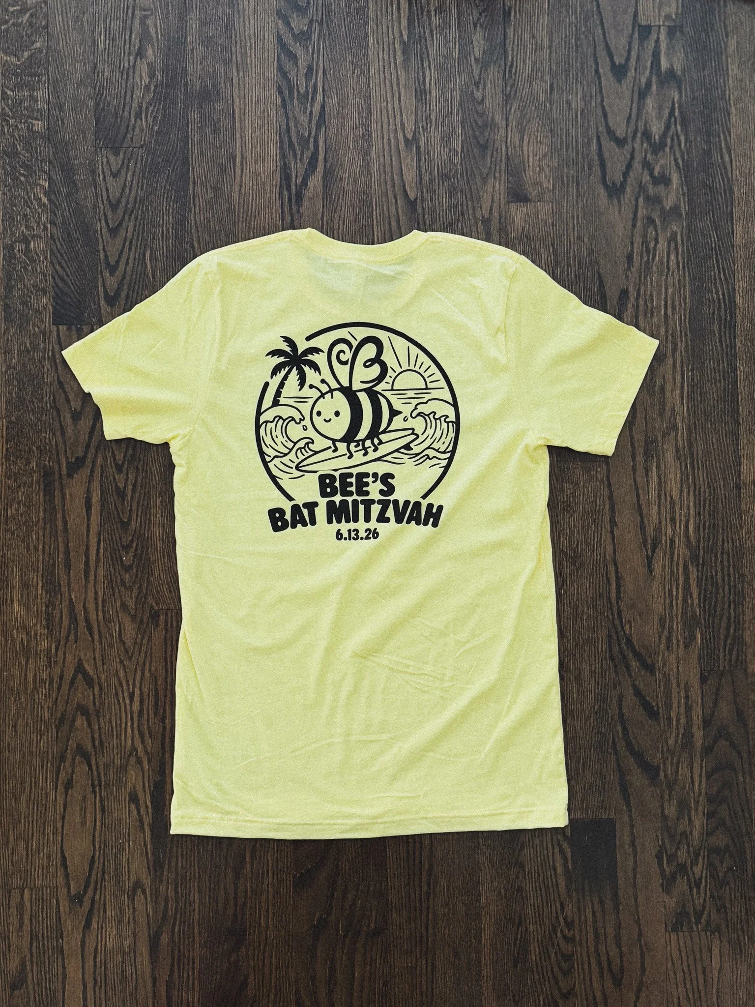 Yellow T-shirt with black graphic design featuring a bee surfing on waves, a palm tree, sun, and the text "Bee's Bat Mitzvah 6.13.26" on a wooden floor.