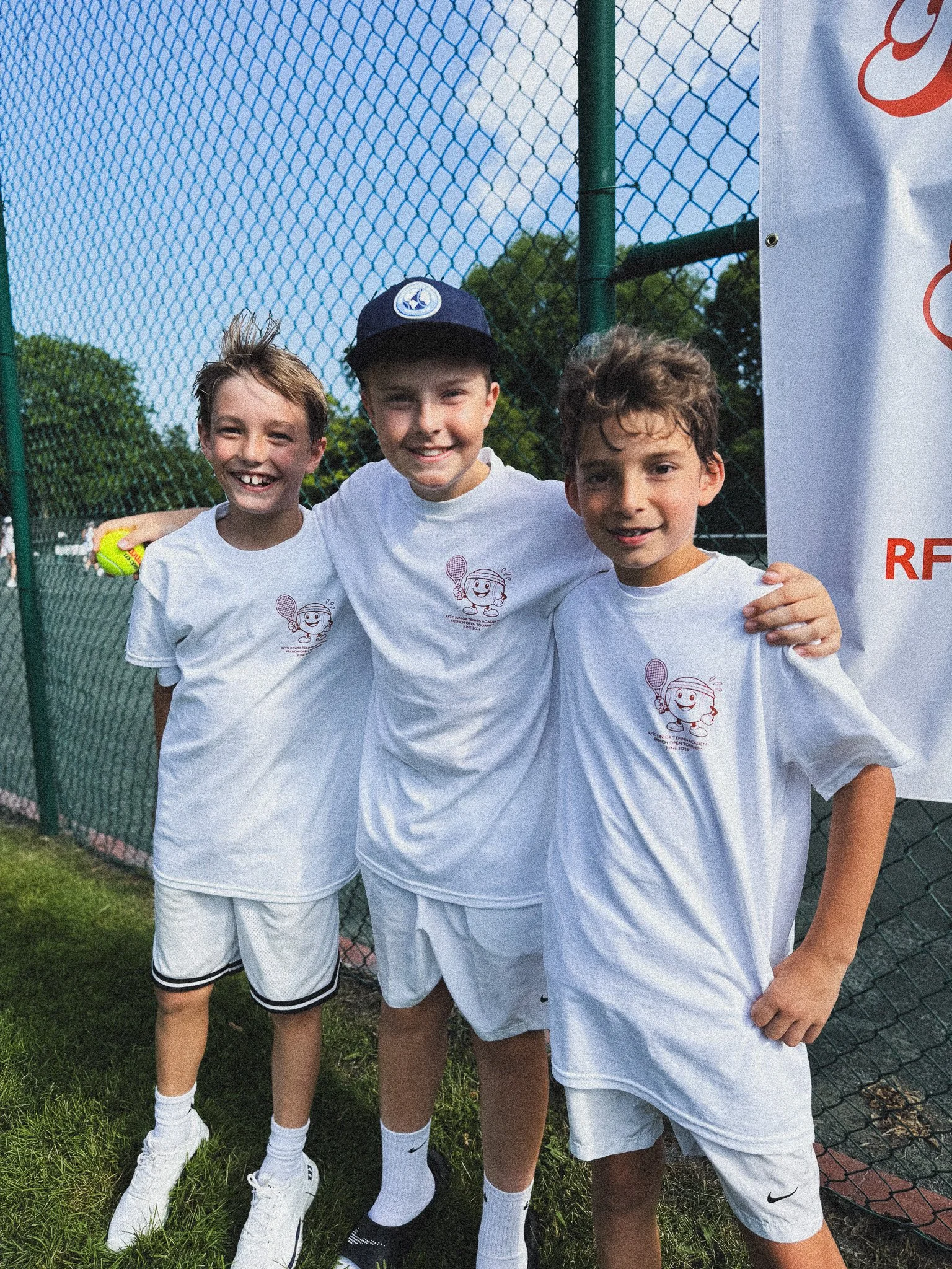 Three young boys wearing white tennis shirts and shorts, standing outdoors near a chain-link fence, smiling for the camera, with trees and a blue sky in the background.