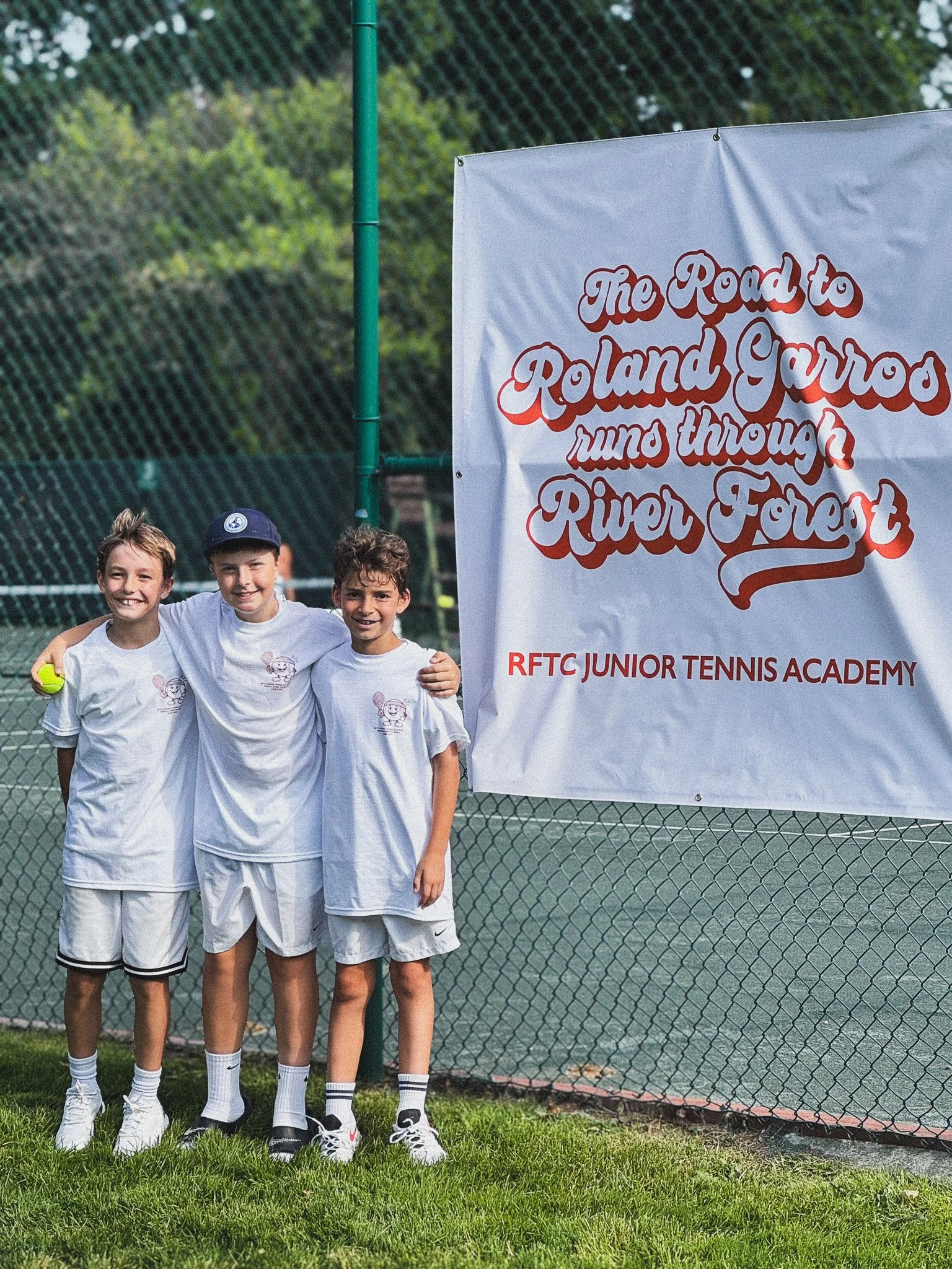 Three young boys in white tennis uniforms and hats standing together on a tennis court with a banner that reads 'The Road to Roland Garros runs through River Forest'.