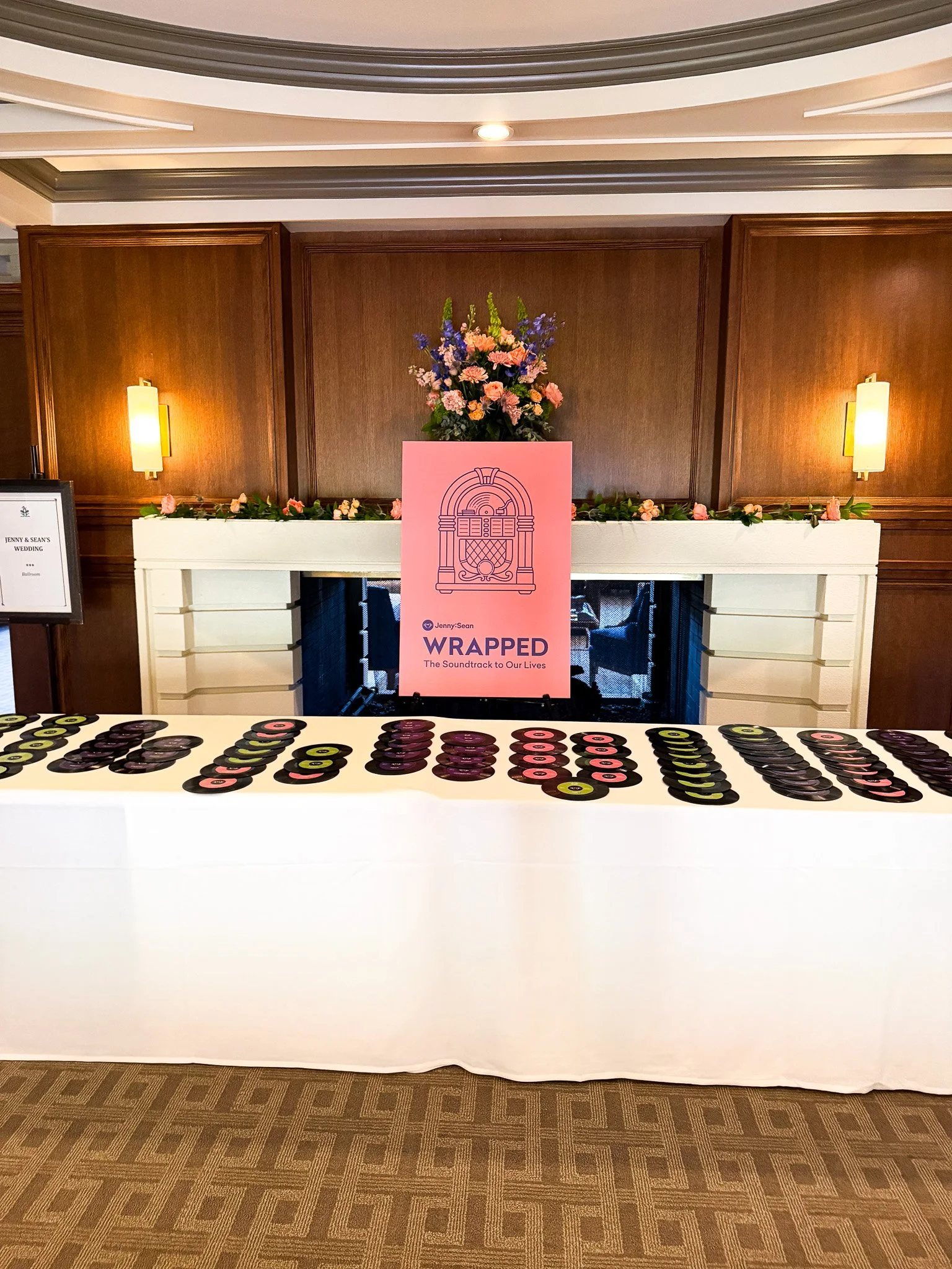 Table with black and pink vinyl records, a pink sign with a line drawing of an arched building and the text 'WRAPPED The Soundtrack to Our Lives', a floral arrangement with pink, purple, and green flowers on a white fireplace mantel.