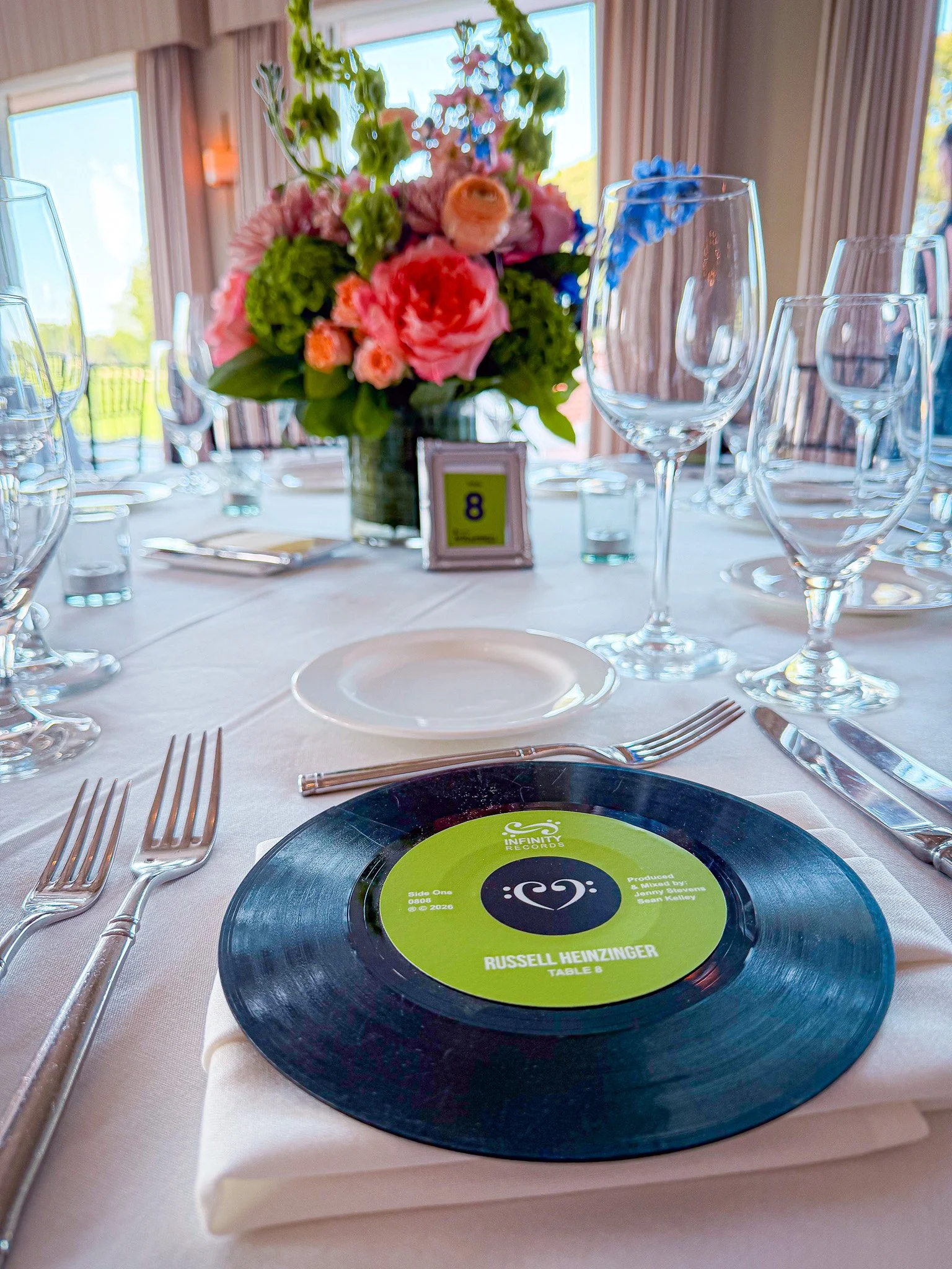 A decorated table with a floral centerpiece, wine glasses, and a vinyl record with a green label stating 'Russell Heizinger, Table 8' at a banquet or event.