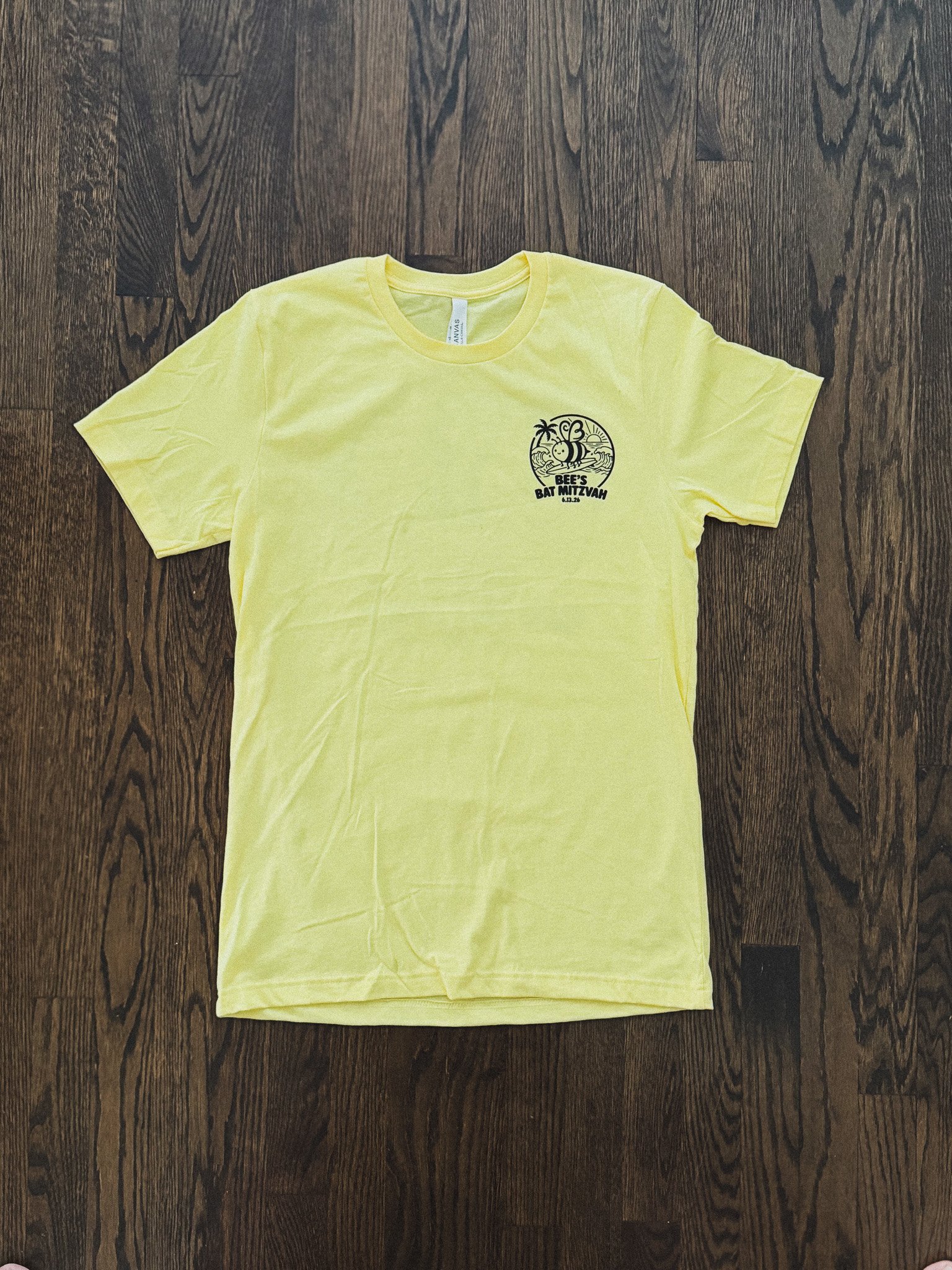 A yellow T-shirt with a black graphic design of a bee, palm trees, and waves, and the text "BEE'S BAT MITZVAH 6.13.26" on the front, laid flat on a dark wooden floor.