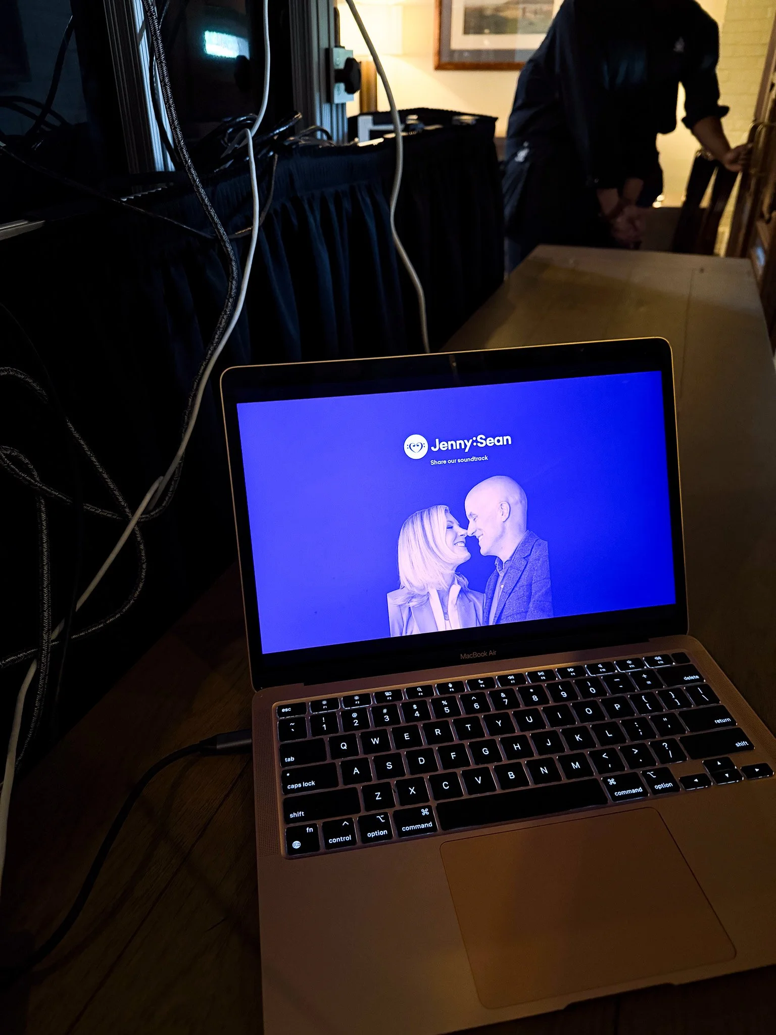A laptop displaying a music playlist titled 'Jenny:Sean' with an image of a couple smiling at each other, set on a wooden table with a man in the background standing near a door.