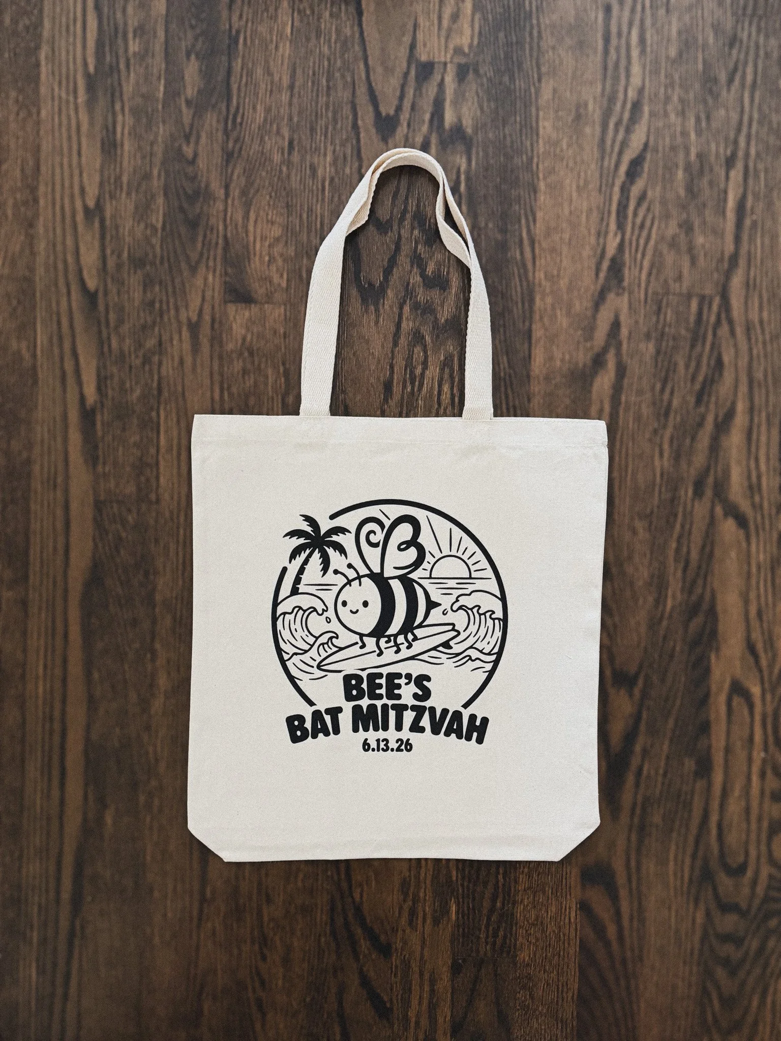 A white tote bag hanging on a wooden wall with a black circular logo featuring a smiling cartoon bee surfing on a wave, palm trees, a sunset, and the text 'BEE'S BAT MITZVAH 6.13.26'.