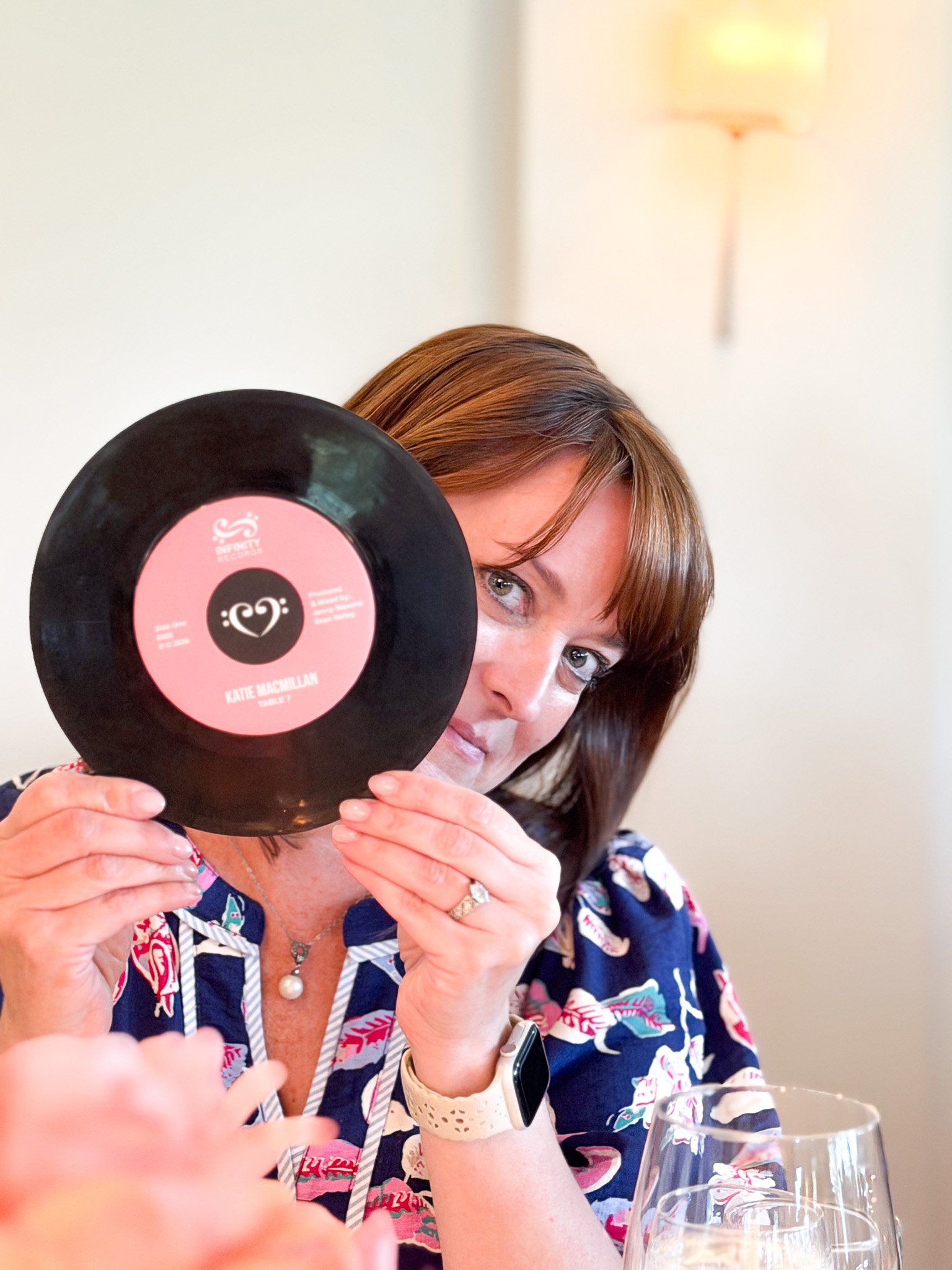 A woman with brown hair holding a black vinyl record with pink label partially covering her face, sitting at a table with wine glasses, and a hand in the foreground.