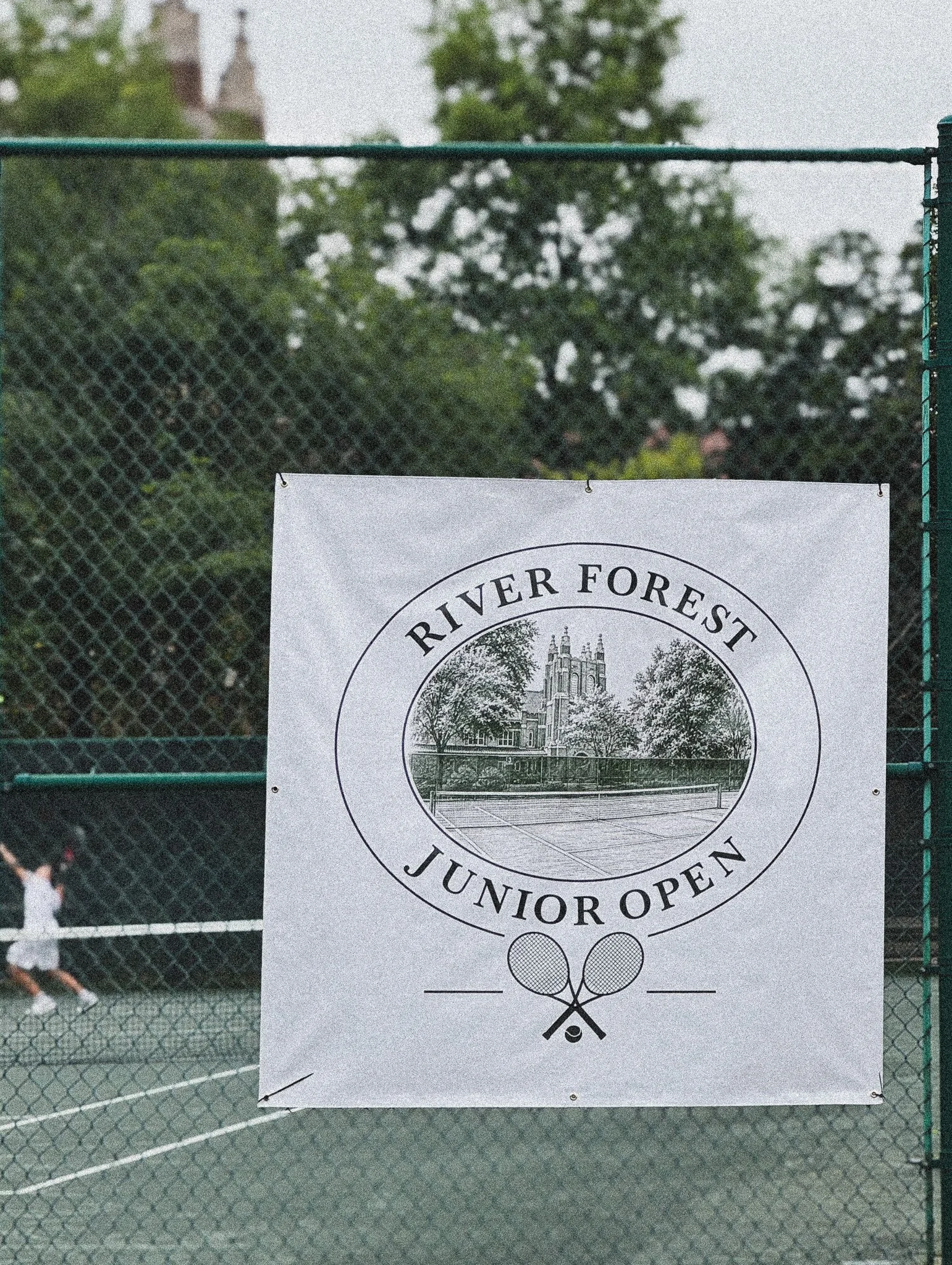 Sign on a tennis court with text 'River Forest Junior Open', a circular logo of a building surrounded by trees, and crossed tennis rackets below.