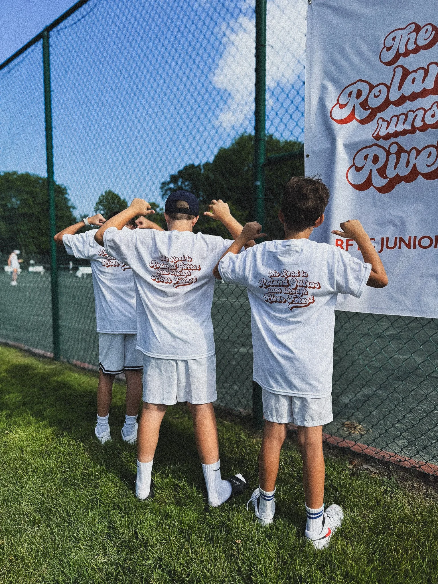 Four young children in white T-shirts and sports shorts standing by a chain-link fence, flexing their arms. The T-shirts have text that reads, 'The Road to Roland Garros runs through River Forest.' They are on a tennis court, outdoors, during daytime.