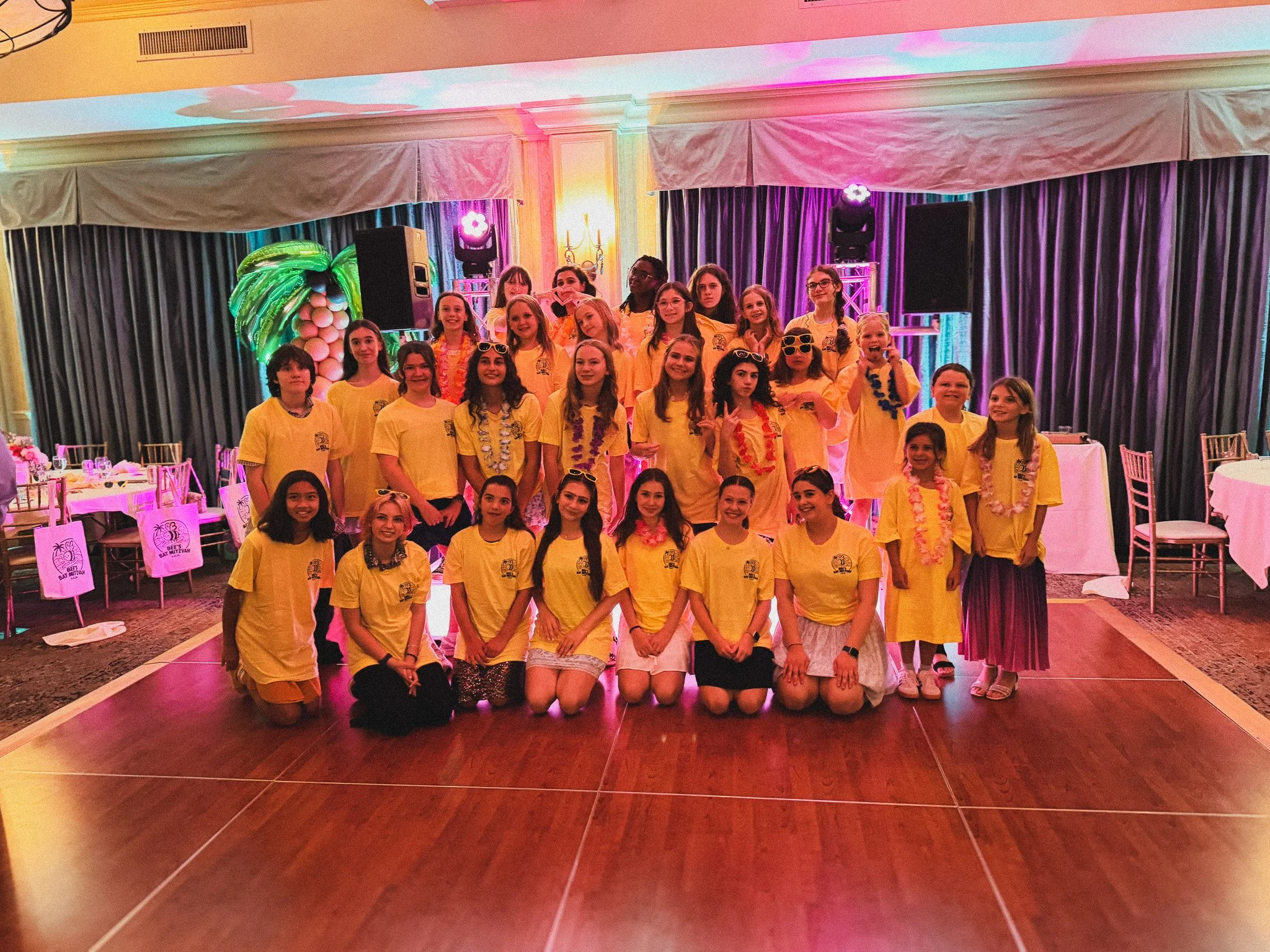 Group of children in yellow shirts at a colorful event with balloons and stage lighting.