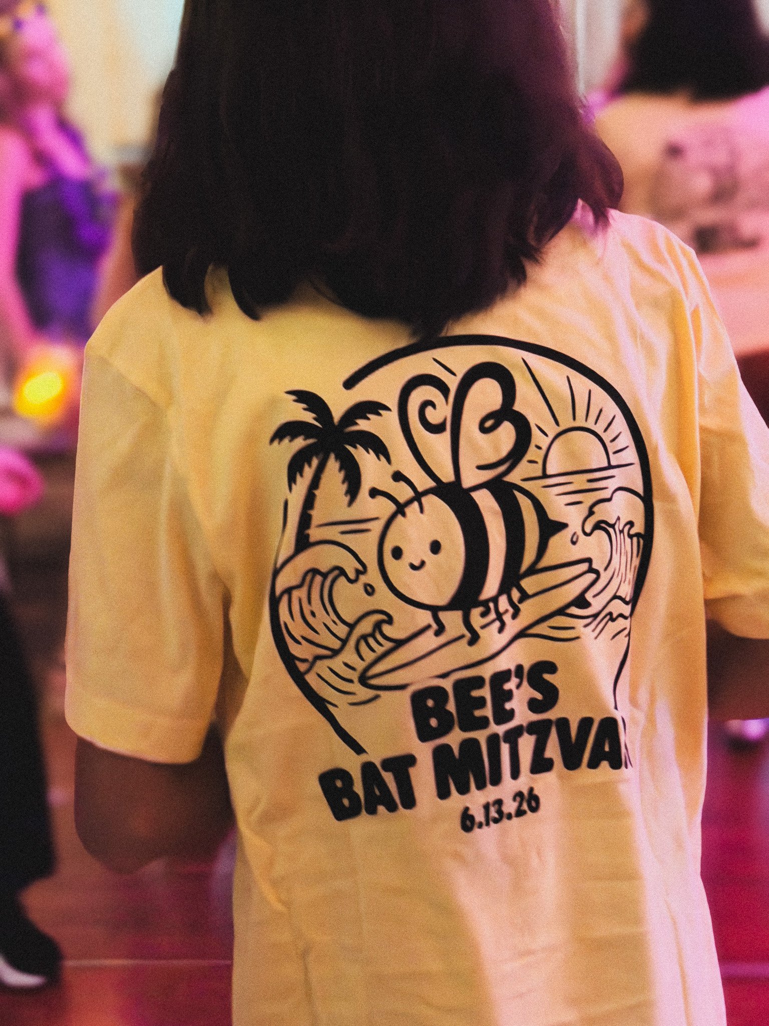 Person wearing a yellow T-shirt with a logo and text promoting the Bat Mitzvah event for Bee's Bat Mitzva, scheduled for June 13, 2026. The logo features a smiling cartoon bee with a palm tree and sunset in the background.