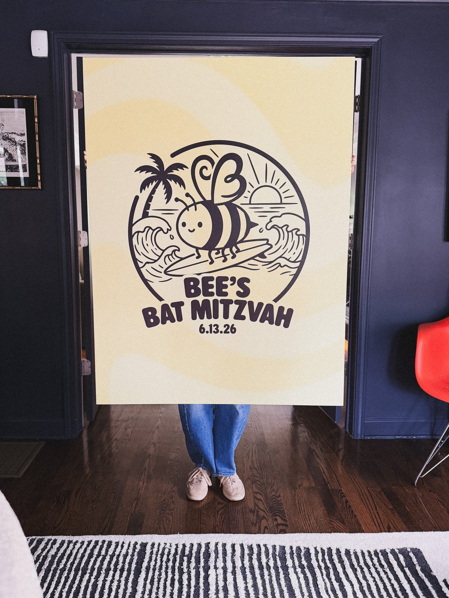 Person holding a large design sign that features a cartoon bee on a surfboard riding the waves at sunset, with the words 'Bee's Bat Mitzvah' and the date 6.13.26.