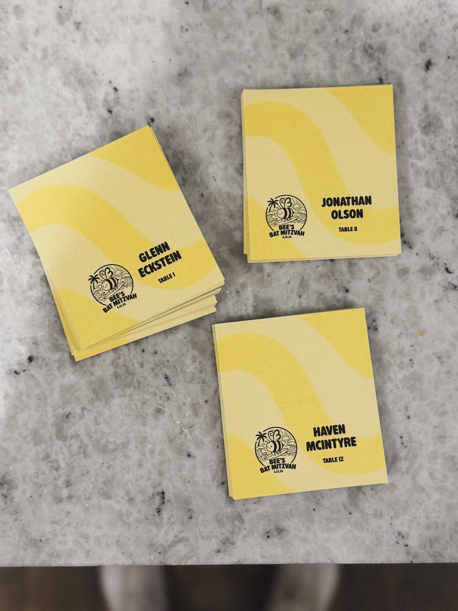Event name badges on a marble surface, each displaying a person's name, table number, and event logo with a bee, palm tree, and sunset, for Bee's Bat Mitzvah.
