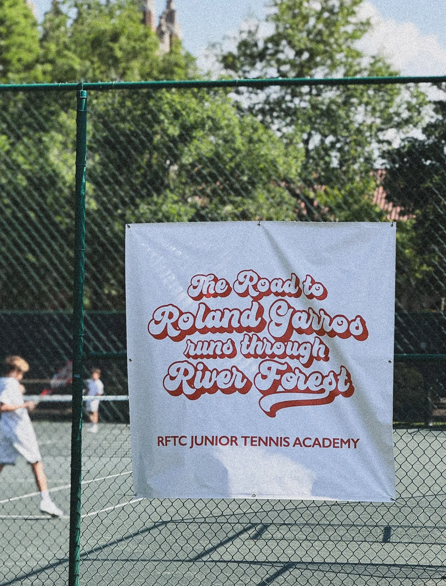 A tennis court with a sign hanging on a chain-link fence. The sign displays the text "The Road to Roland Garros, RFTC Junior Tennis Academy." Some children are visible in the background, walking near the court, with trees and a cloudy sky overhead.
