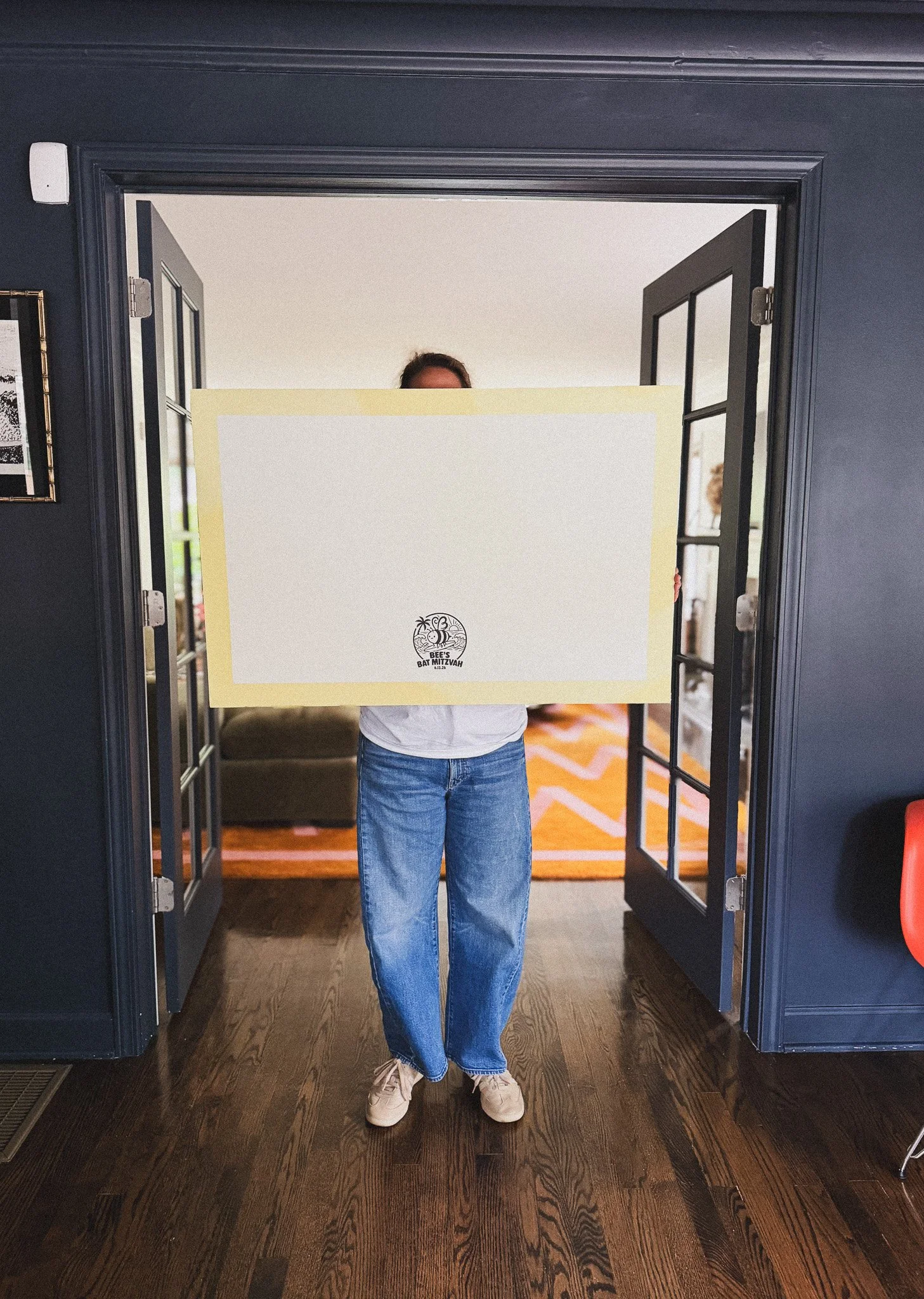 Person standing in a doorway holding a large white sign in front of their face, with only their forehead visible, wearing jeans and sneakers, in a room with dark walls and hardwood floor.