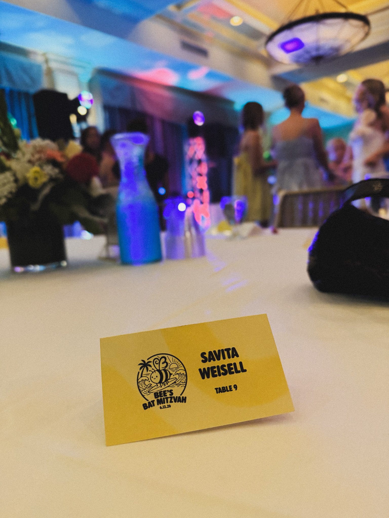 A yellow table number card with black writing indicating it is for Beé's Bat Mitzvah, dated 4.13.20, for table 9, placed on a white table. In the blurred background, there are people standing and talking at an event, with decorative lighting and floral arrangements visible.