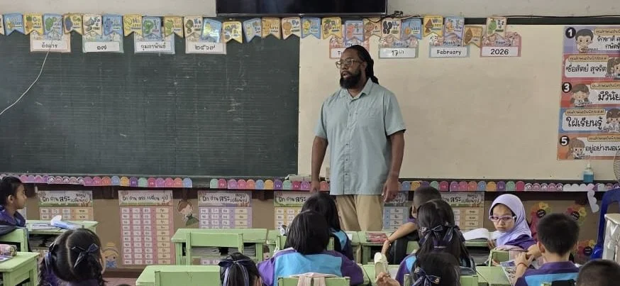 A classroom with young students seated at green desks, facing a teacher standing near a chalkboard. The chalkboard has colorful banners and educational posters above it, with some text in a different alphabet and numbers, including the year 2026. The students are paying attention to the teacher, who is wearing glasses and a light blue shirt.