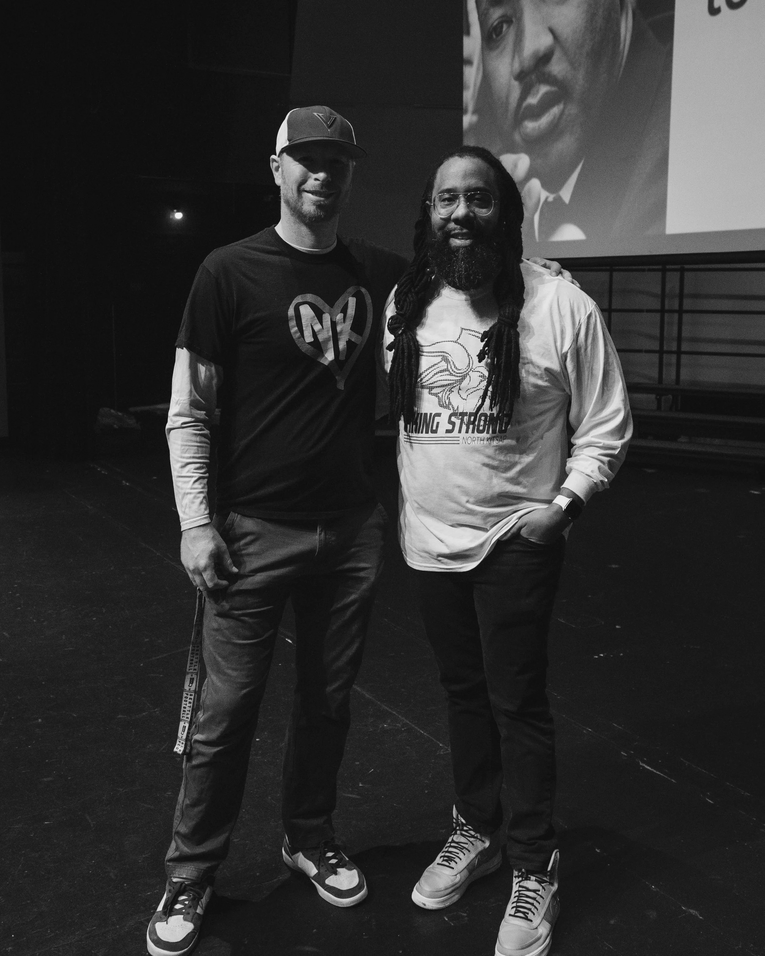 Two men standing together in a dimly lit room, smiling at the camera. One man wears a baseball cap and a t-shirt, the other has glasses, a beard, and dreadlocks, wearing a long-sleeve shirt with a graphic design. A large black-and-white portrait of Martin Luther King Jr. is projected on the wall behind them.