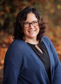 A woman with curly dark hair and glasses smiling outdoors with autumn foliage in the background, wearing a blue cardigan over a black top.