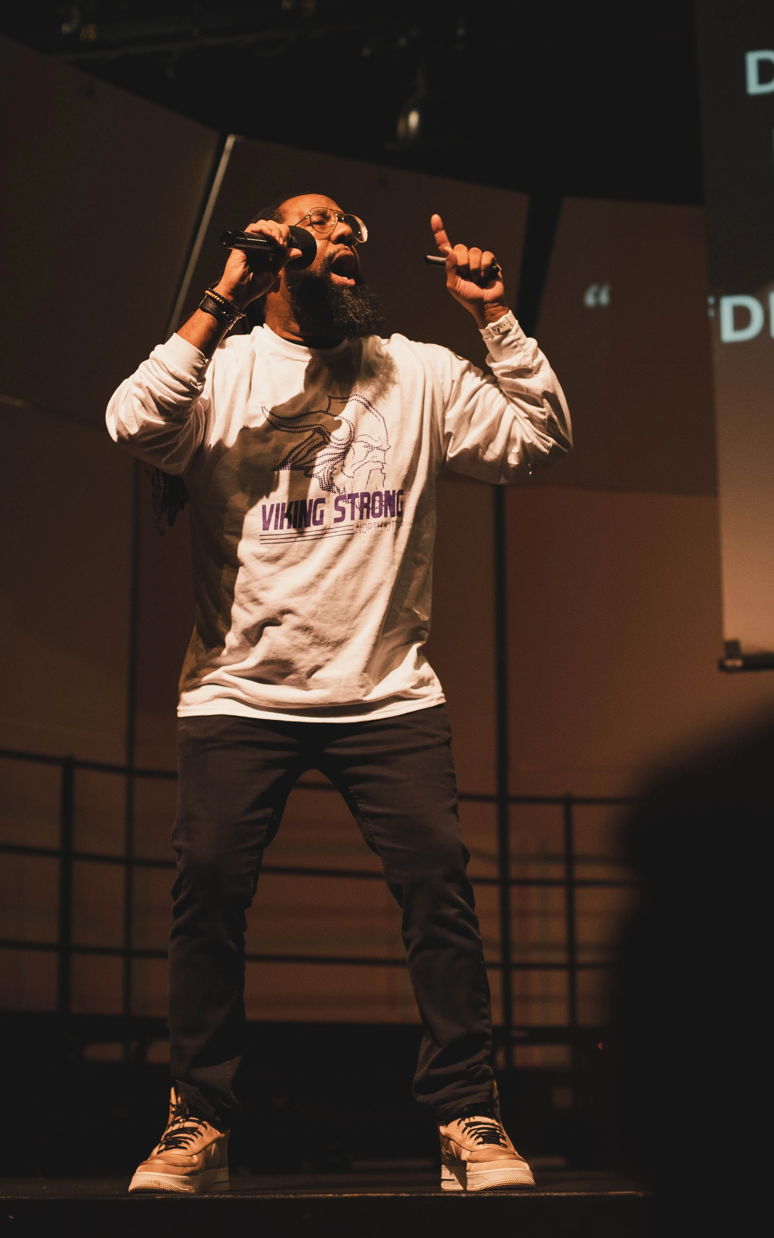 A man with a beard and glasses singing into a microphone on stage, wearing a white sweatshirt and sneakers, with his right hand raised and index finger pointing upward.