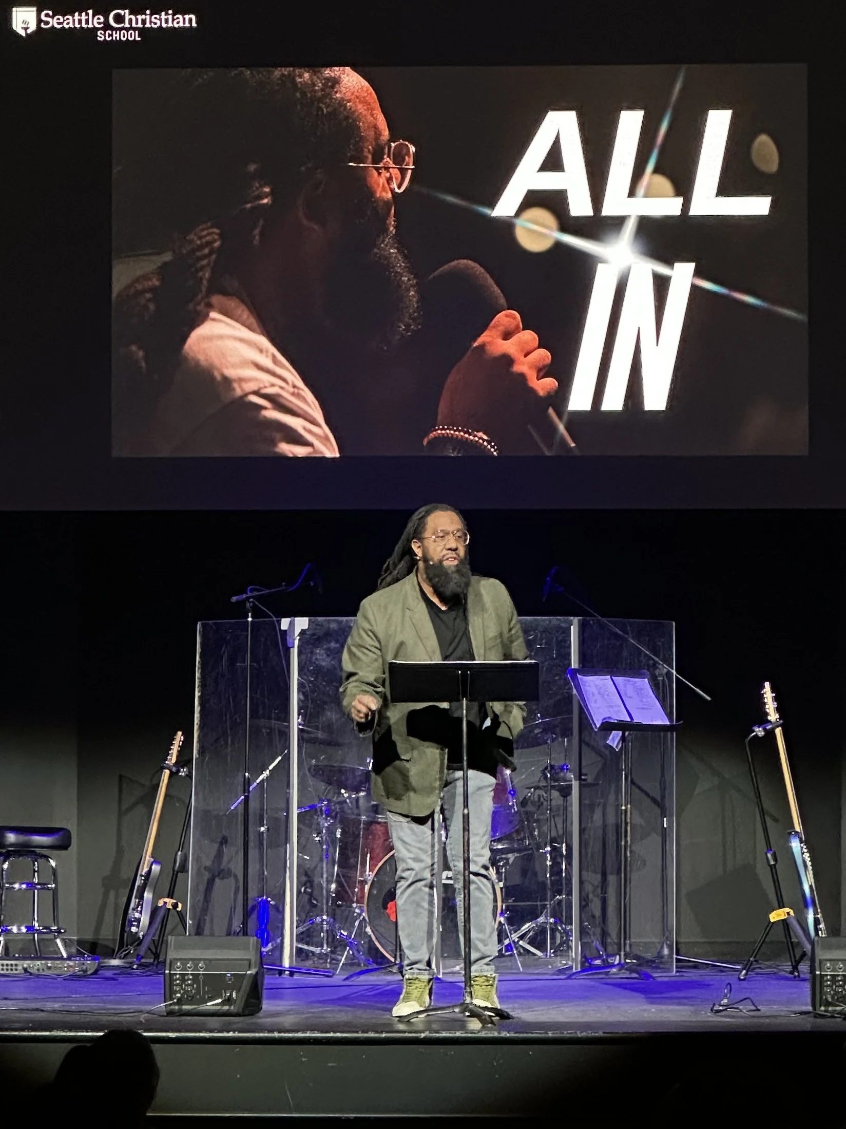 A man with long hair and a beard, wearing glasses, a green blazer, and yellow shoes, stands at a microphone on a stage with musical instruments behind him. A large screen above displays an image of another man with glasses and dreadlocks, with the words "ALL IN" in large white letters. The logo for Seattle Christian School is in the top left corner.