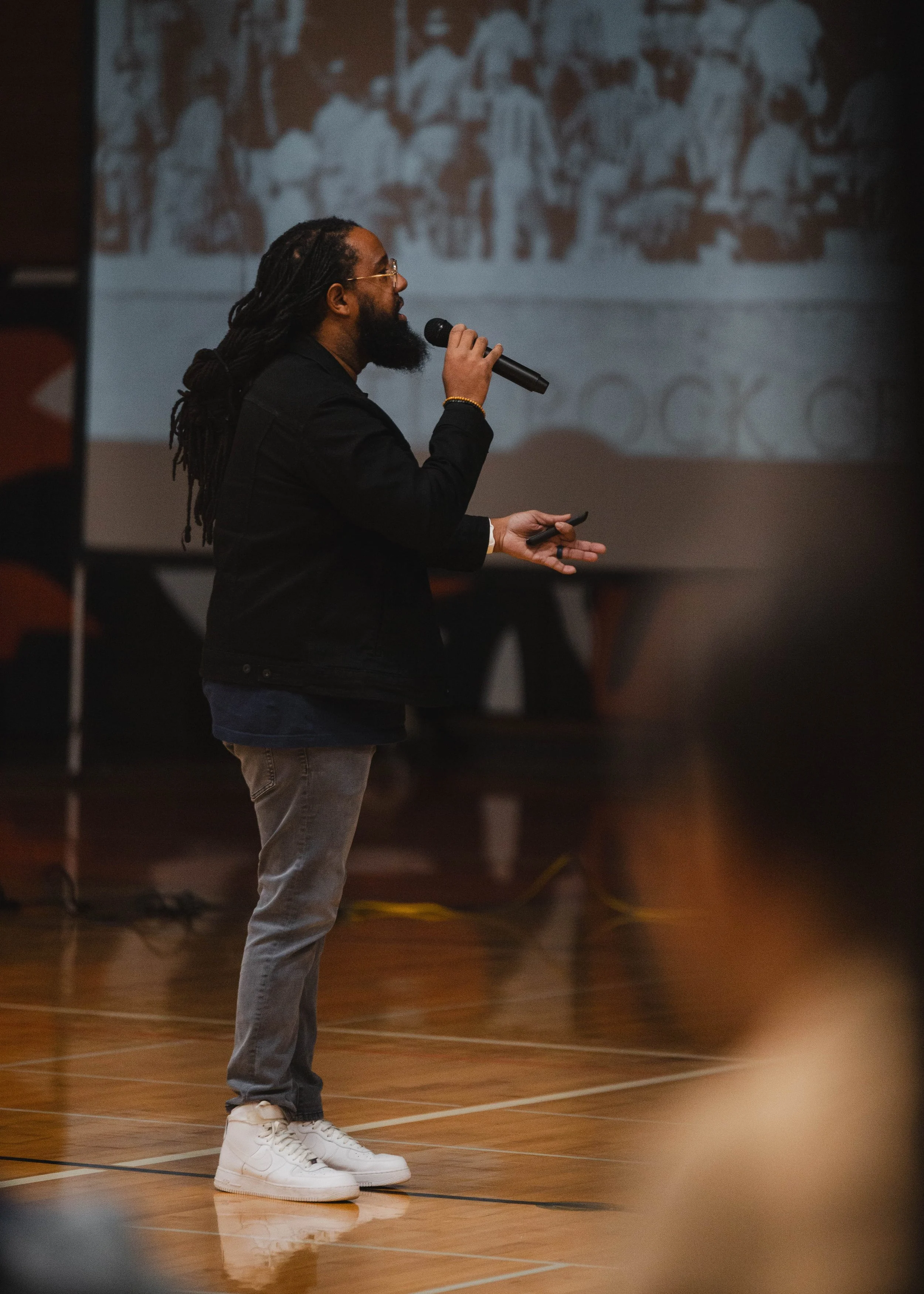A person with long dreadlocks and glasses holding a microphone, speaking at an indoor event with a projected presentation in the background.