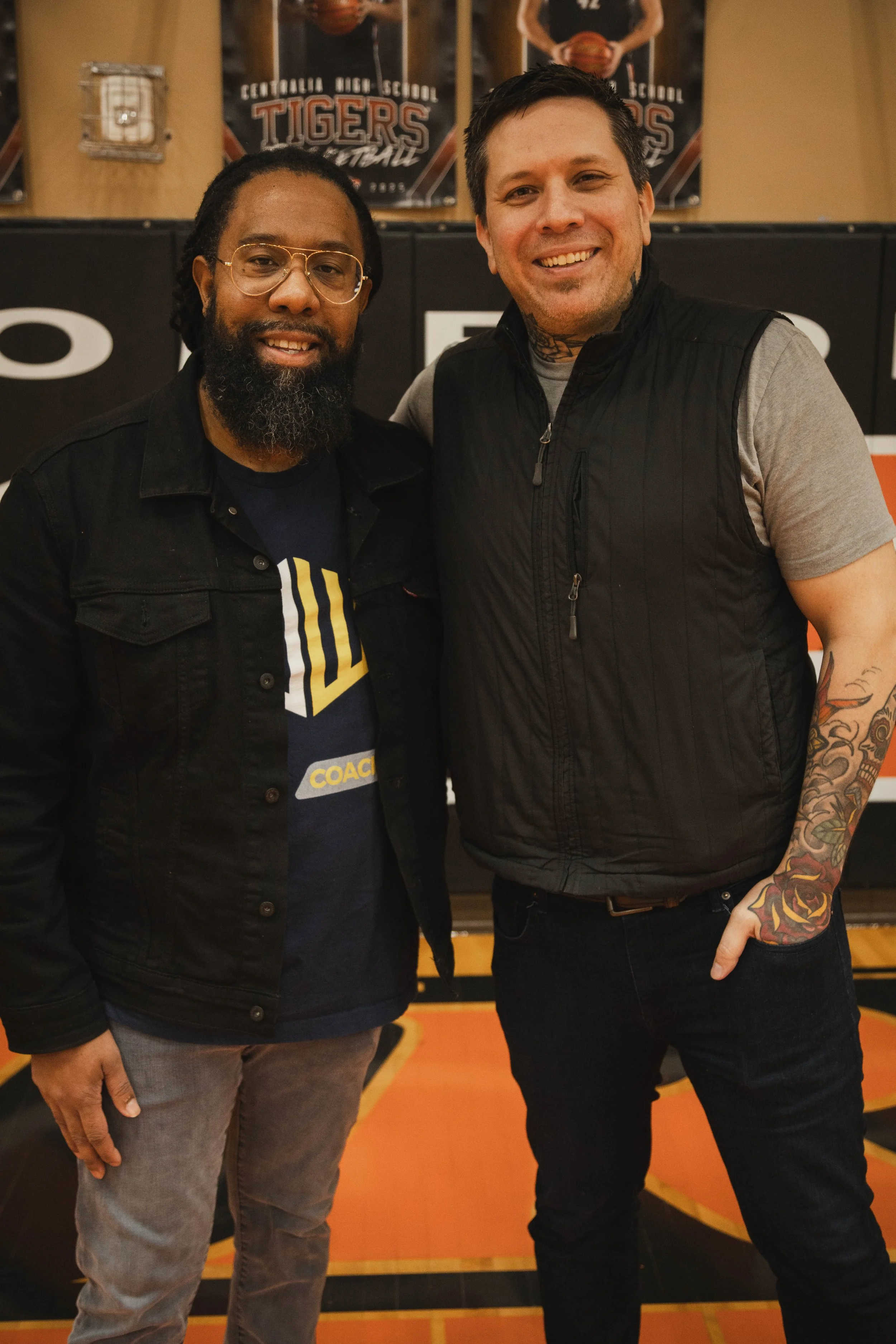 Two men standing on a basketball court, smiling at the camera. The man on the left has long dreadlocks, glasses, a beard, and is wearing a black jacket over a shirt with a design. The man on the right has short dark hair, tattoos on his neck and arms, and is wearing a gray t-shirt with a black vest. In the background, there are posters for Centalia West School Tigers.