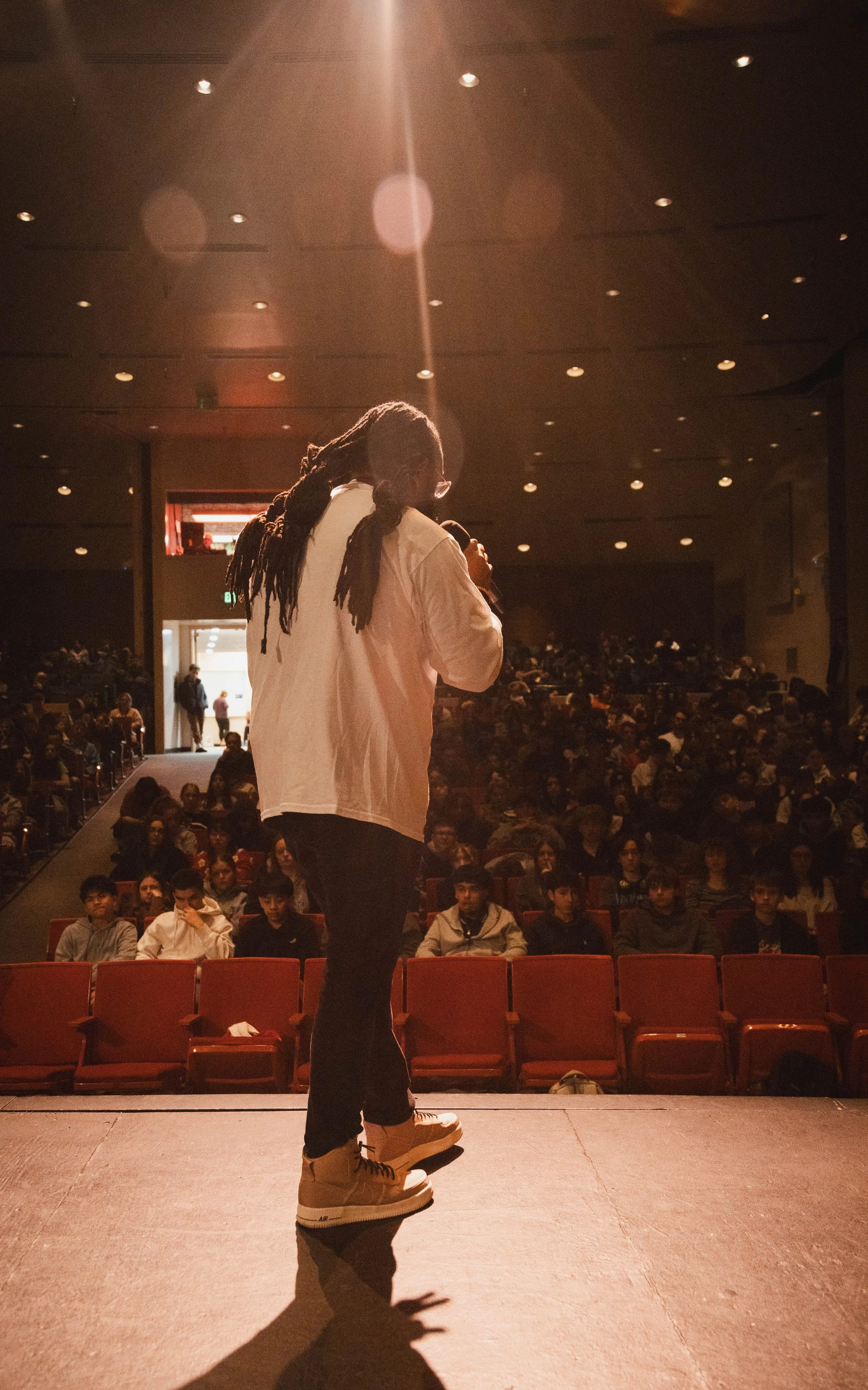 A man with dreadlocks and glasses speaking into a microphone on stage in front of an audience in a theater with padded seats and a high ceiling.