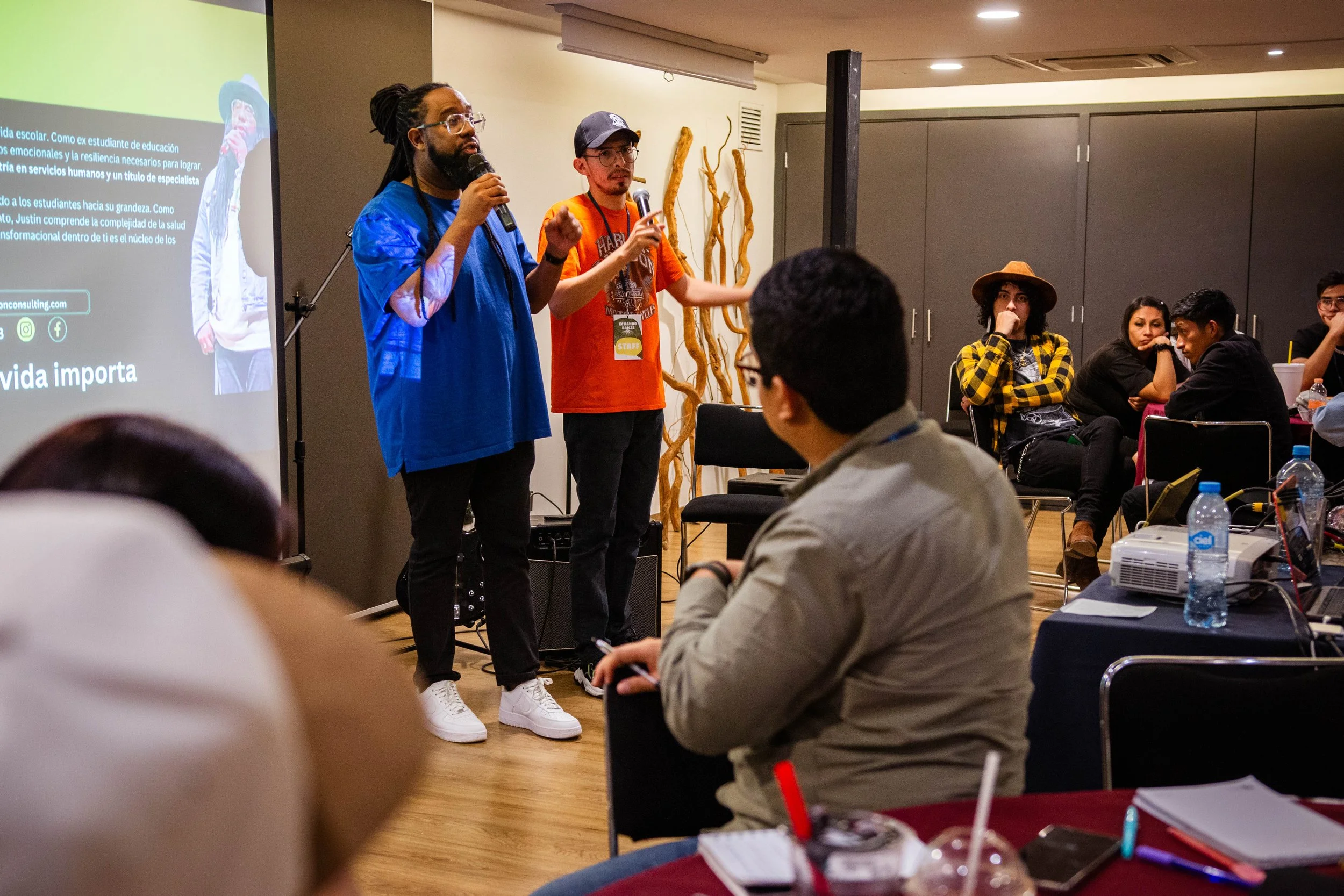 Two men are standing and speaking into microphones in front of an audience during a presentation or workshop. The man on the left has dreadlocks, glasses, a beard, and is wearing a blue jacket. The man on the right has short hair, glasses, and is wea