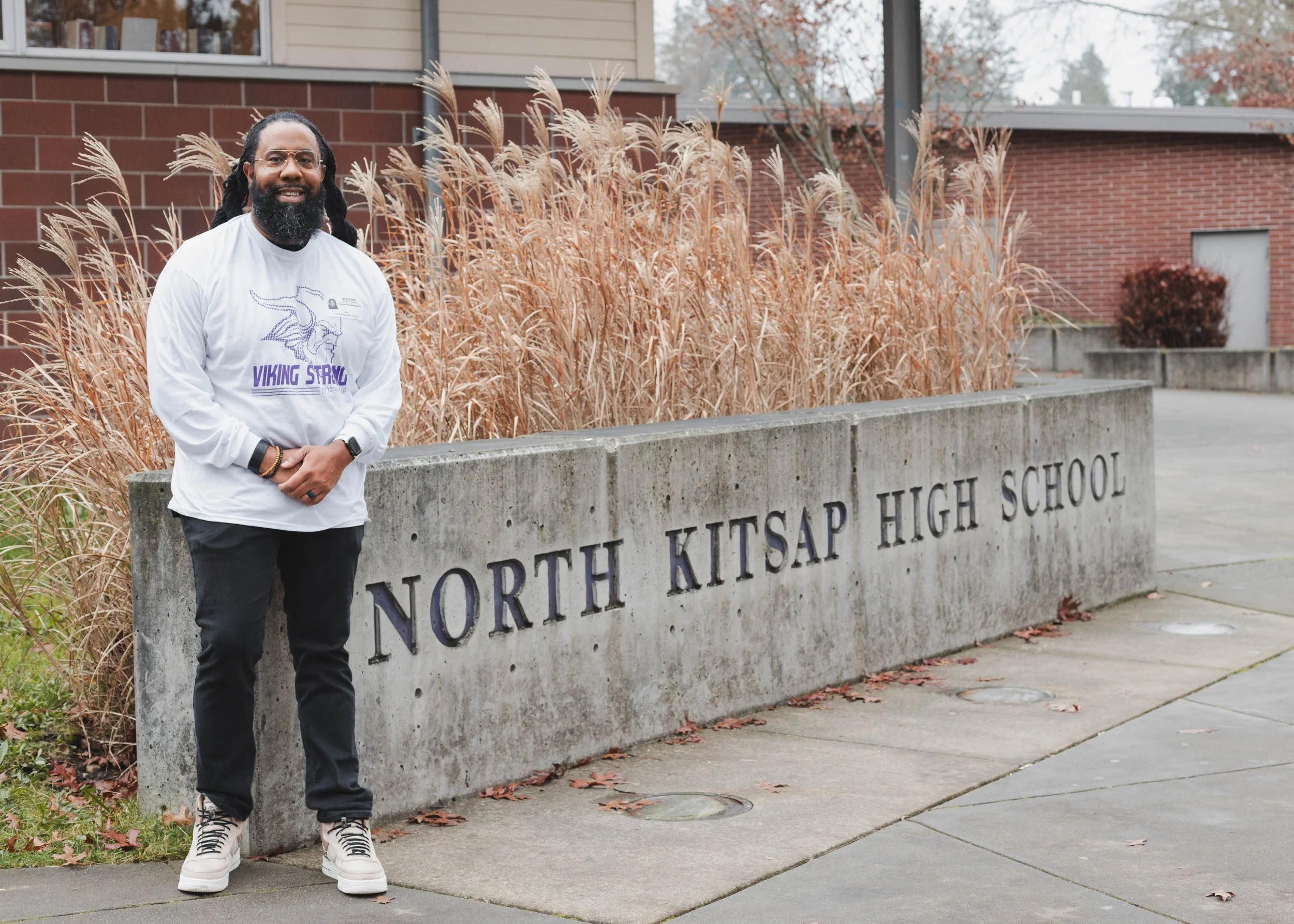 A man with glasses, a beard, and dreadlocks standing outdoors next to a concrete sign that reads "North Kitsap High School." He is wearing a white shirt with a Viking logo and the words "Viking Strong," black pants, and white sneakers. The background features a brick building, tall dry grass, and trees with autumn leaves.