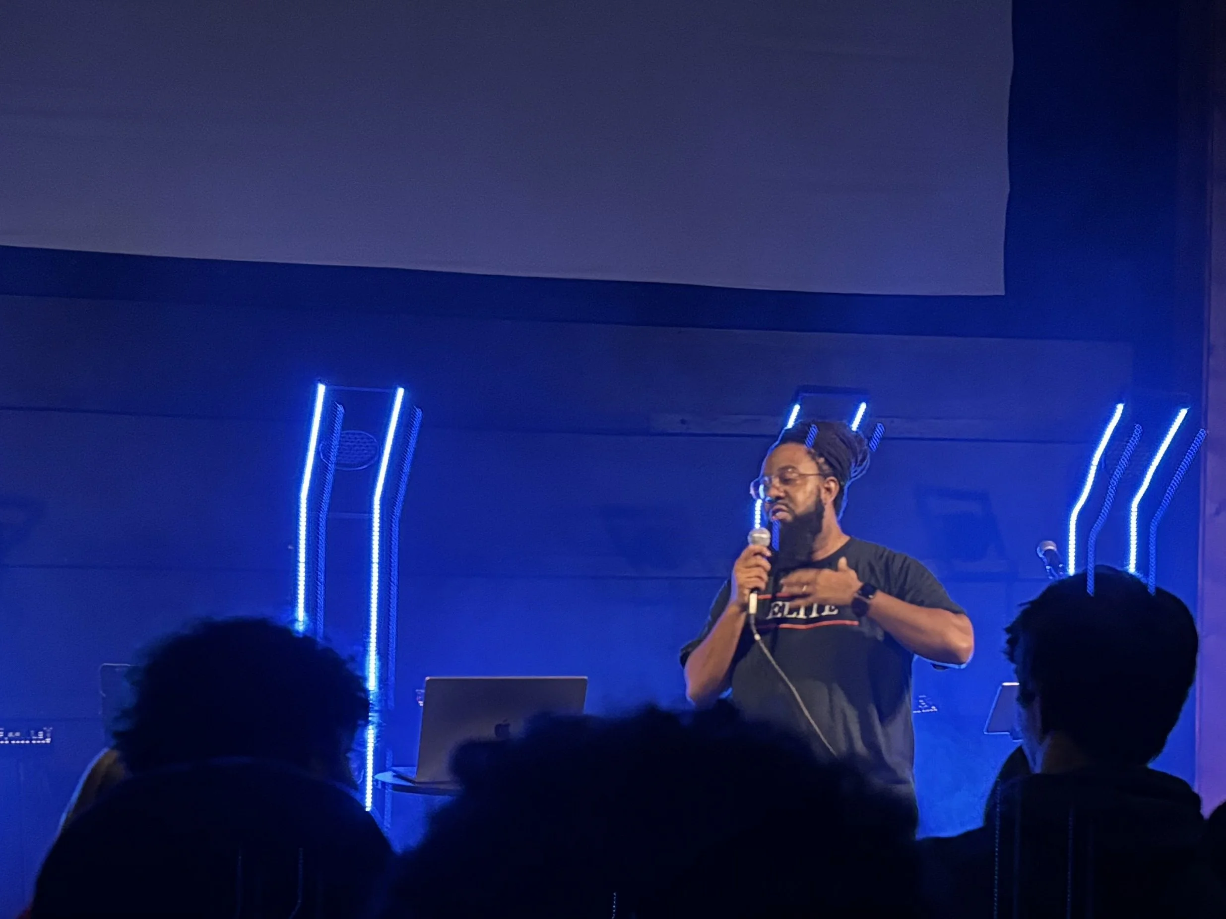 A man with glasses and a beard, holding a microphone, speaking on stage with blue vertical light strips behind him in a dark room. Audience silhouettes are visible at the bottom.