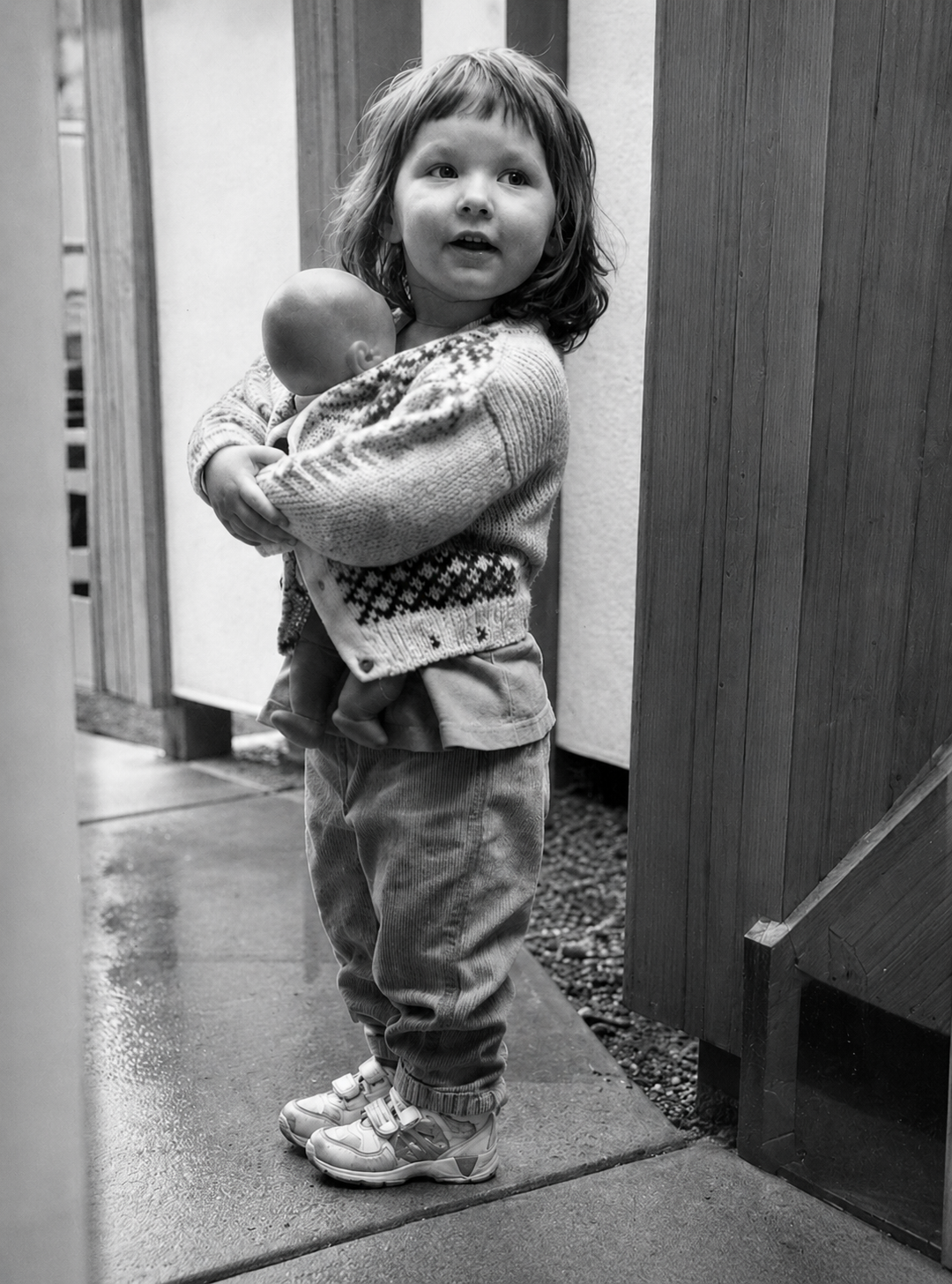 A young girl with curly hair holding a baby, standing outdoors near wooden structures, in black and white.