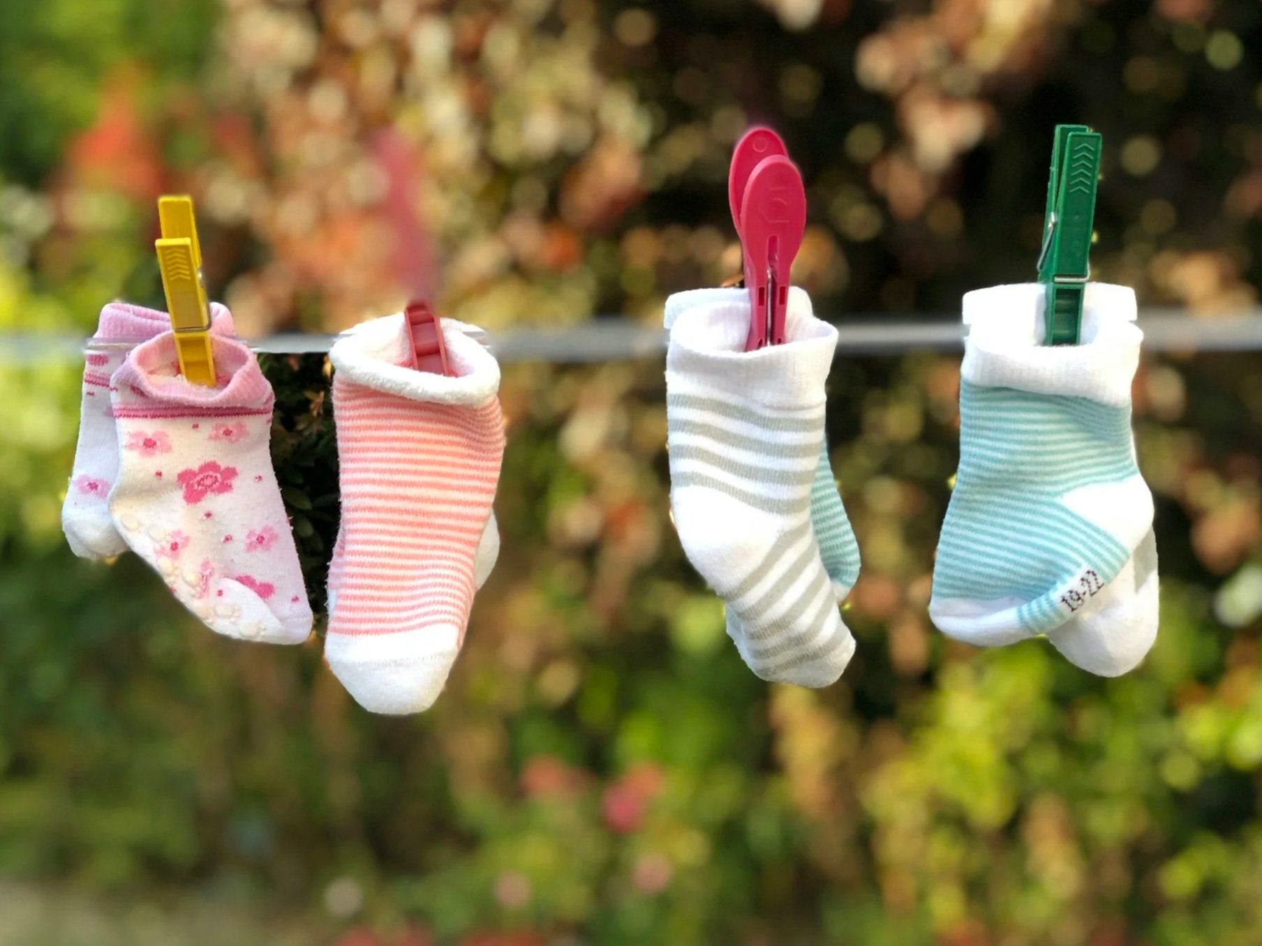 Four baby socks hanging on a clothesline with clothespins, with a blurred garden background.