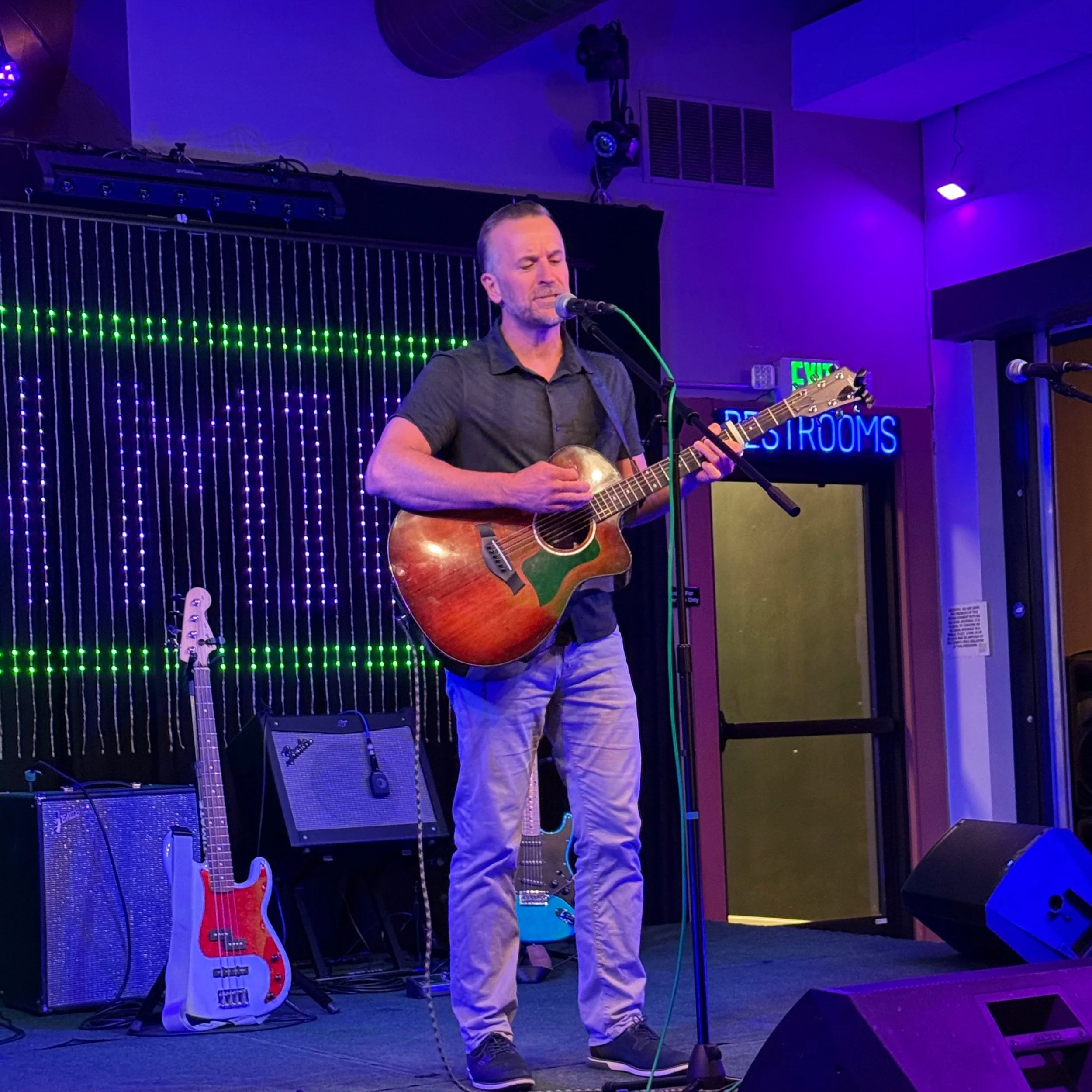 Adam Hill playing guitar on stage with purple and blue lighting, two guitars on stands behind him, illuminated exit sign, and a small amplifier.