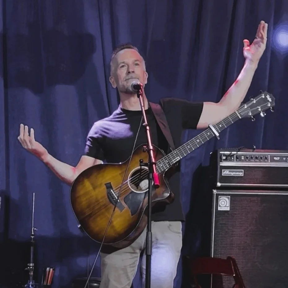 Adam Hill wearing a black T-shirt and beige pants, standing on stage with a sunburst acoustic guitar, raising one arm, and speaking or singing into a microphone. There is a large amplifier behind him and dark curtains in the background.