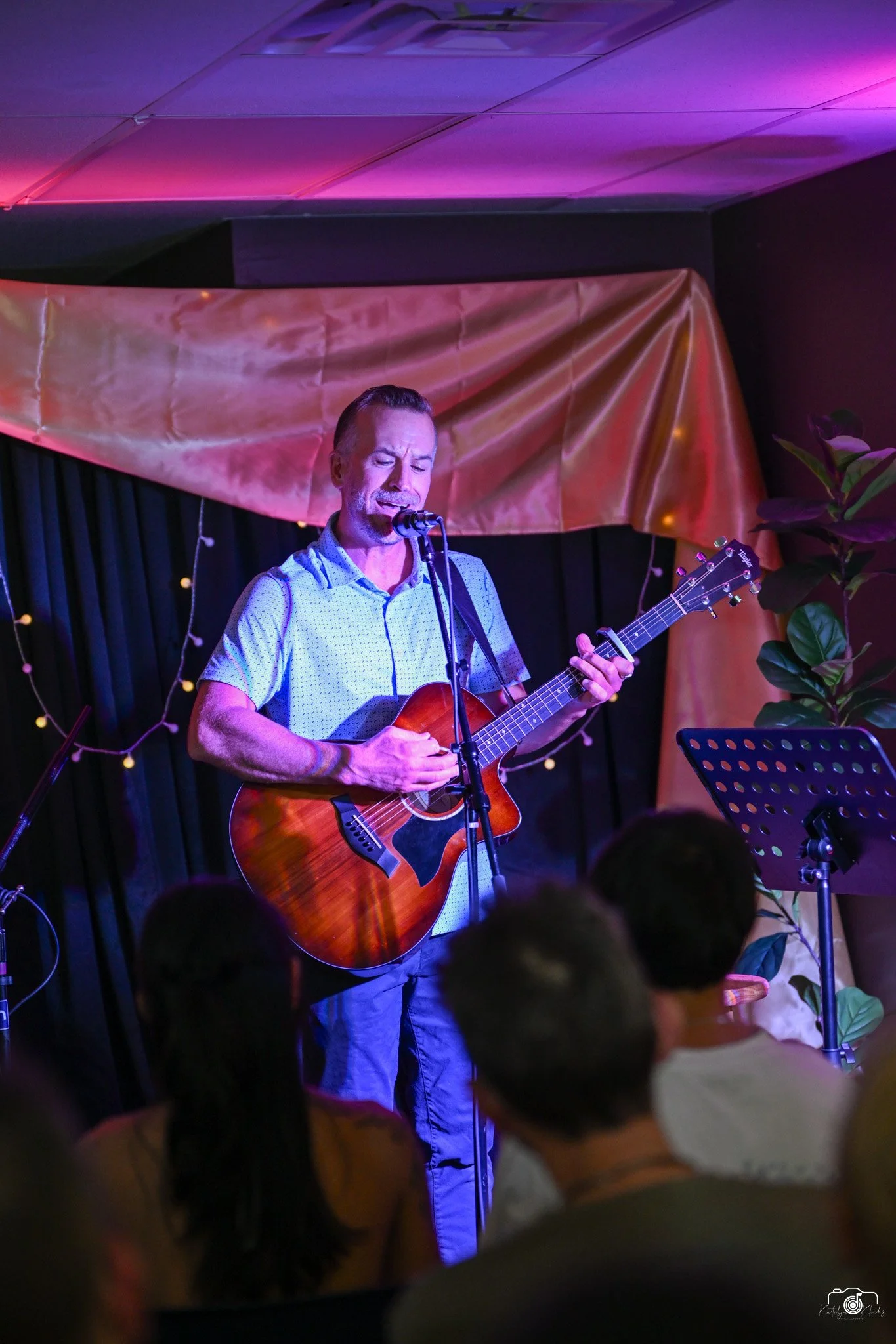 Adam Hill playing an acoustic guitar Americana folk rock music on stage in a dimly lit venue with black curtains and a purple and gold backdrop, visible from the audience perspective.