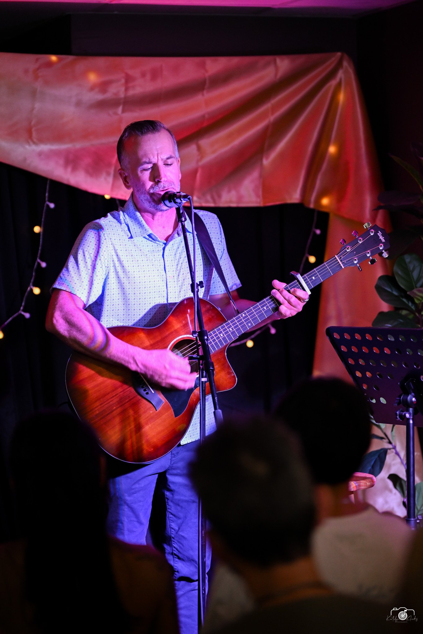 Adam Hill in a light-colored shirt is singing into a microphone and playing an acoustic guitar on stage. The background features a large reddish-orange draped fabric and string lights, creating a warm, intimate performance setting.